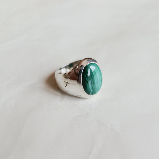 A silver ring featuring an oval green malachite stone, resting on a light beige textured surface.
