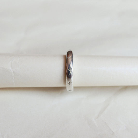 A slim silver band ring with engraved floral details, displayed on a white cylindrical ring stand against a soft, textured off-white background.
