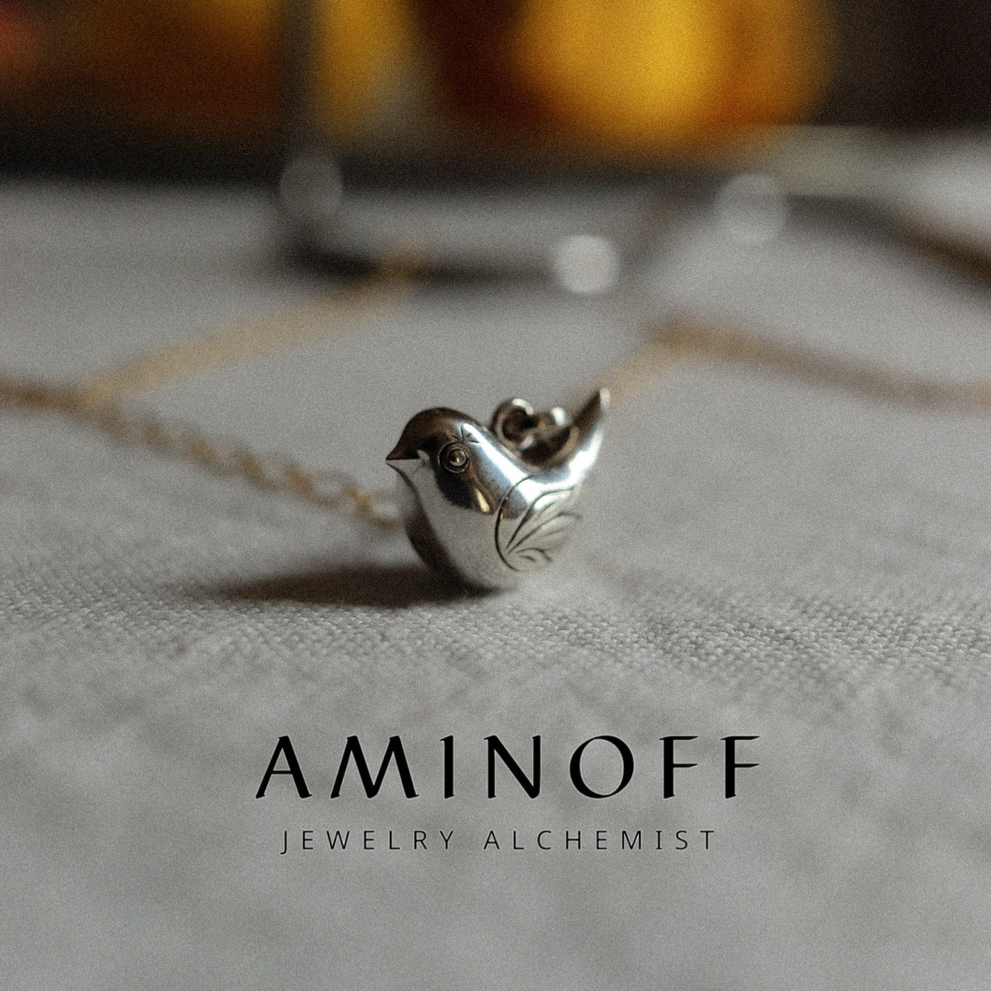 Silver bird-shaped pendant on a chain with 'Aminoff' branding on a textured surface.