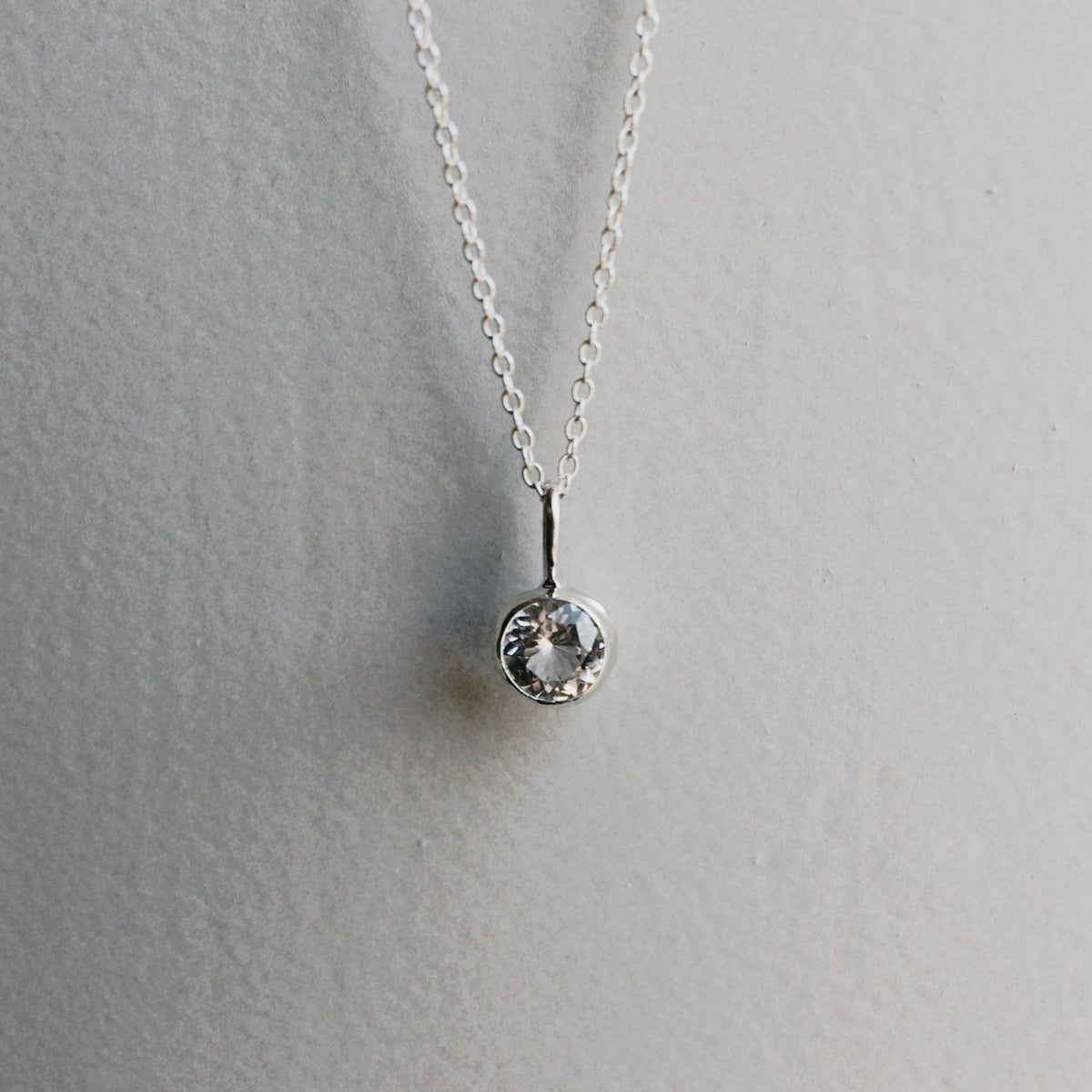 A delicate silver chain necklace with a single bezel-set clear round gemstone pendant, displayed against a neutral light gray textured background.