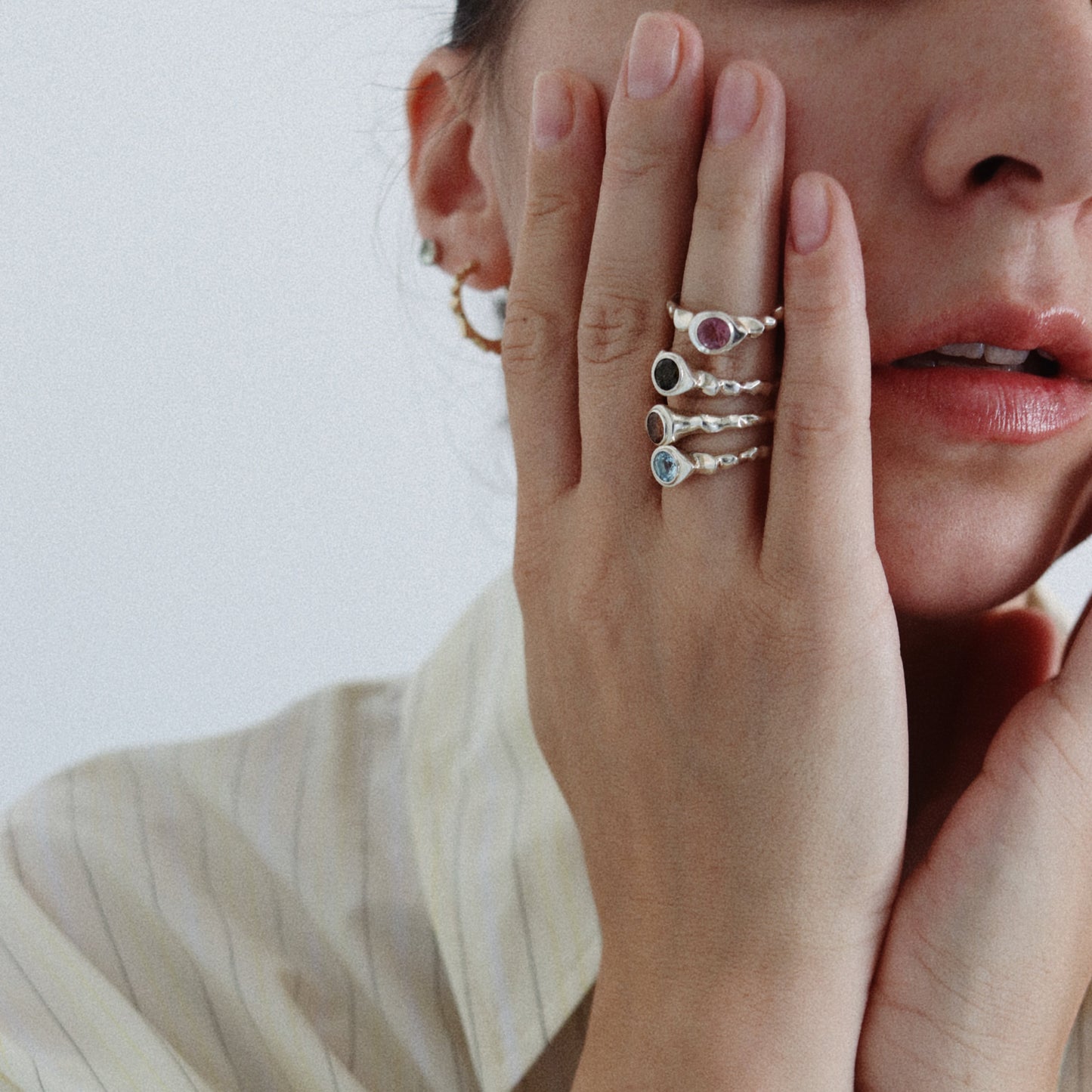 A close-up lifestyle shot of a model wearing three delicate silver rings with small colorful gemstones on her fingers, paired with a cream-colored pinstriped shirt.