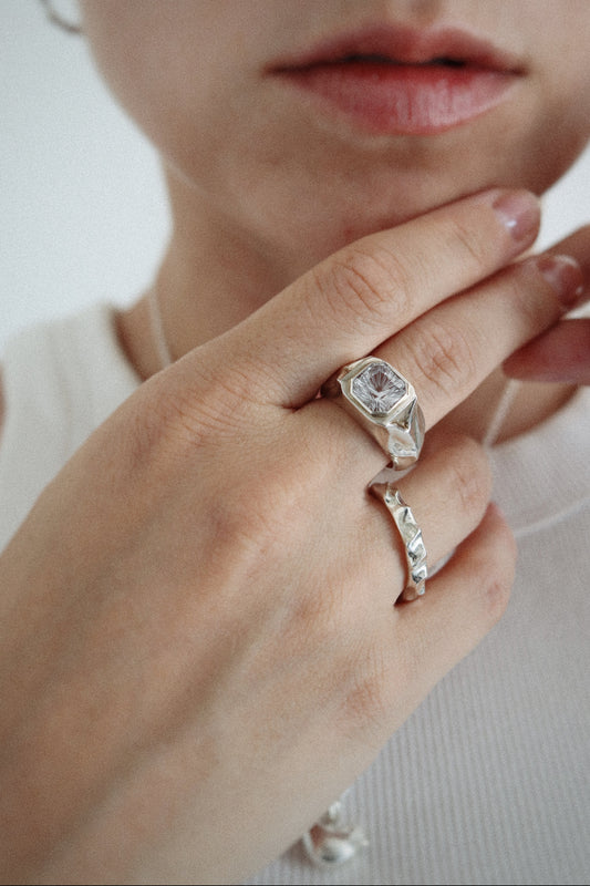 A close-up lifestyle shot of a person wearing a silver signet ring with a clear rectangular gemstone on their middle finger, with another silver ring on their ring finger, set against a clean white background.