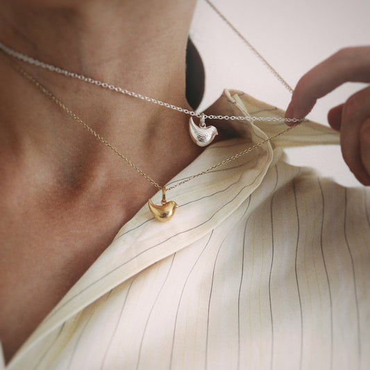 A close-up lifestyle shot of a model wearing two delicate necklaces: a silver chain with a polished silver heart pendant and a gold chain with a gold heart pendant, styled over a cream-colored pinstriped shirt.