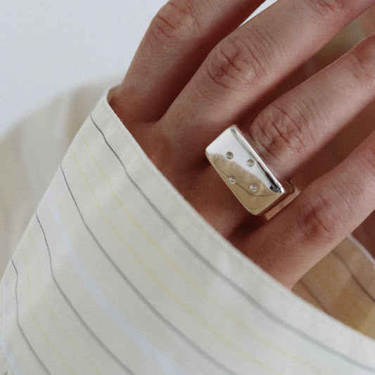 A close-up of a person's hand wearing a chunky, polished gold ring with small clear stones, styled with a cream-colored striped shirt.