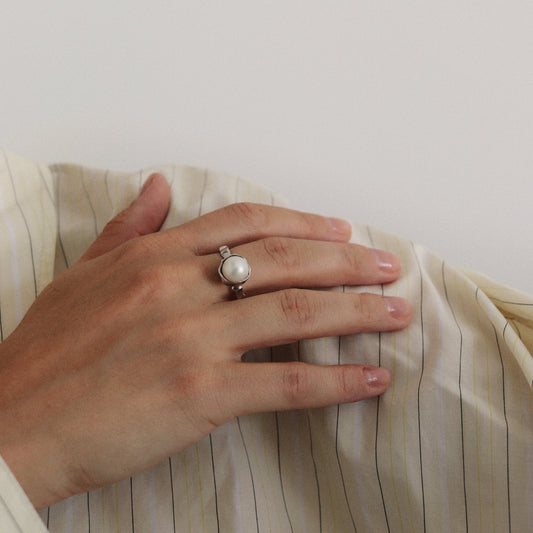 A silver ring with a large white pearl is worn on a person's ring finger, resting on a cream-colored pinstriped fabric against a plain light background.