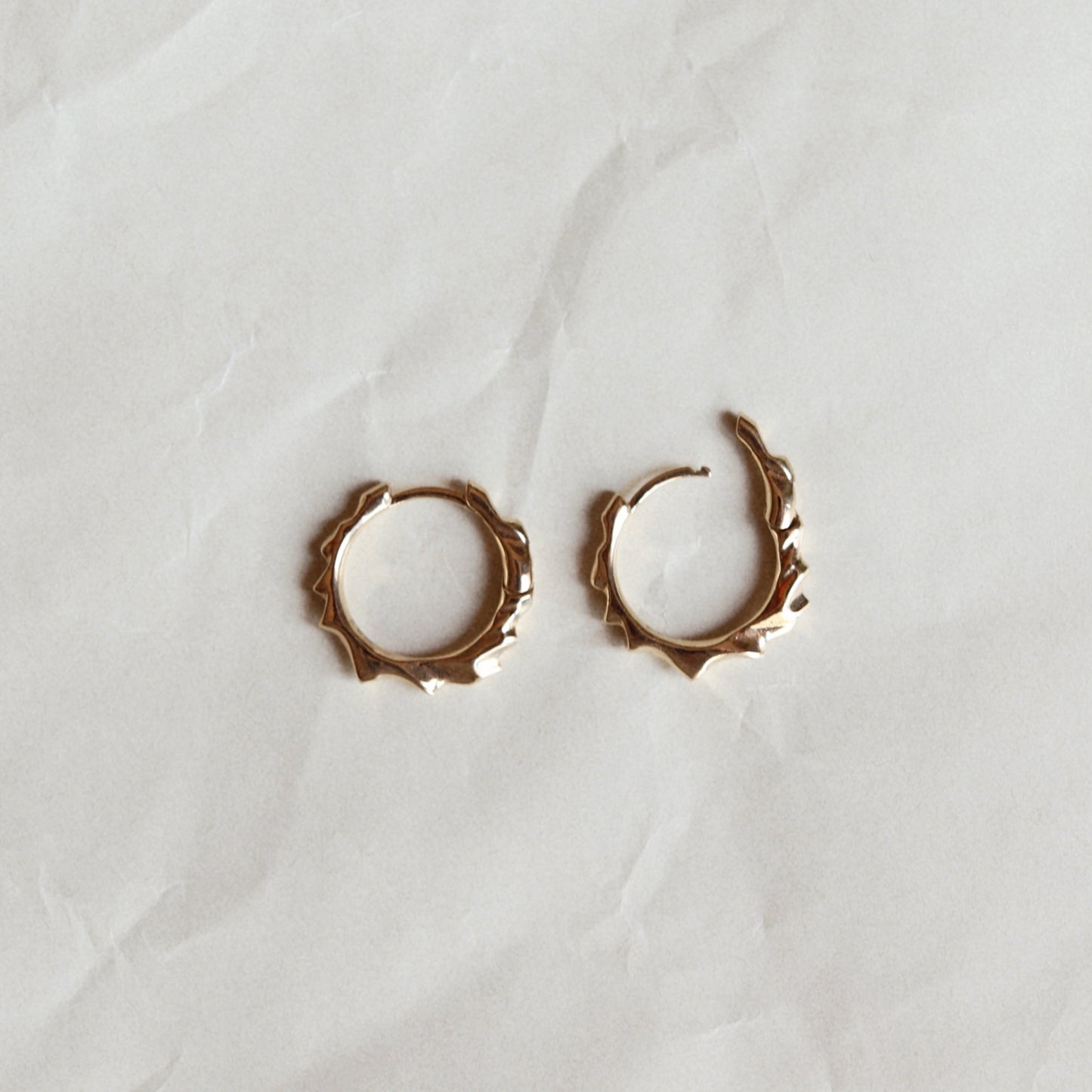 A pair of small gold hoop earrings with a textured, organic jagged edge, displayed on a wrinkled white paper background.