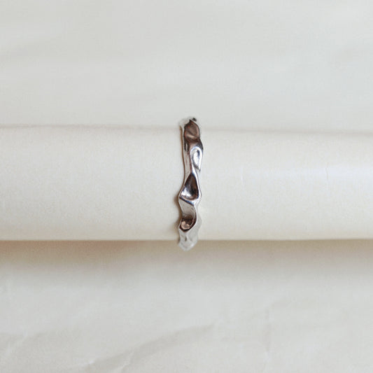 A slim, wavy sterling silver ring displayed on a cream-colored ring holder against a light beige background.