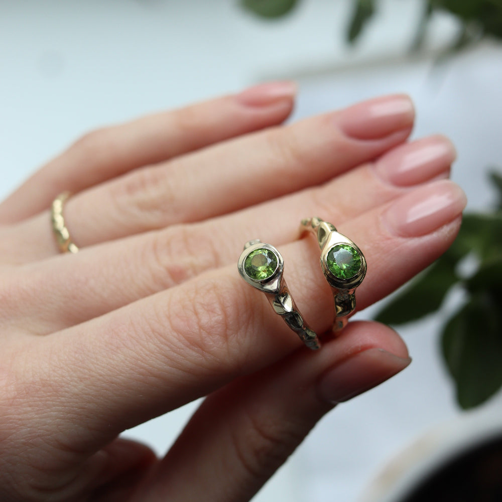 Close-up of a hand wearing two gold rings with green gemstones against a blurred background.