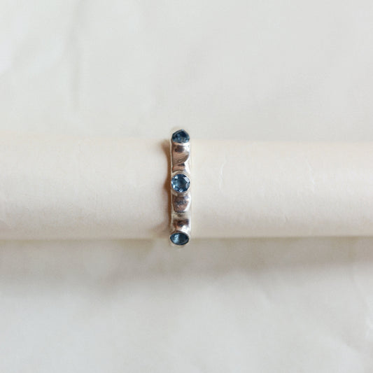 A delicate ring with small blue gemstones and a central clear stone, displayed on a cream-colored ring stand against a soft, light background.