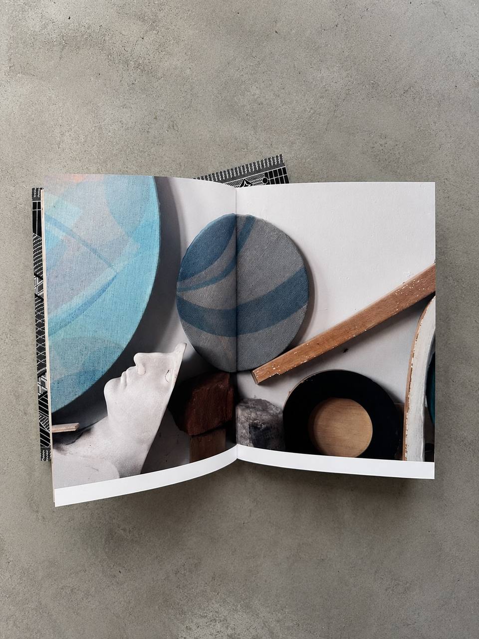An open printed book or magazine laid flat on a textured gray surface, showing an abstract interior photograph with blue circular forms, white space, and neutral objects.
