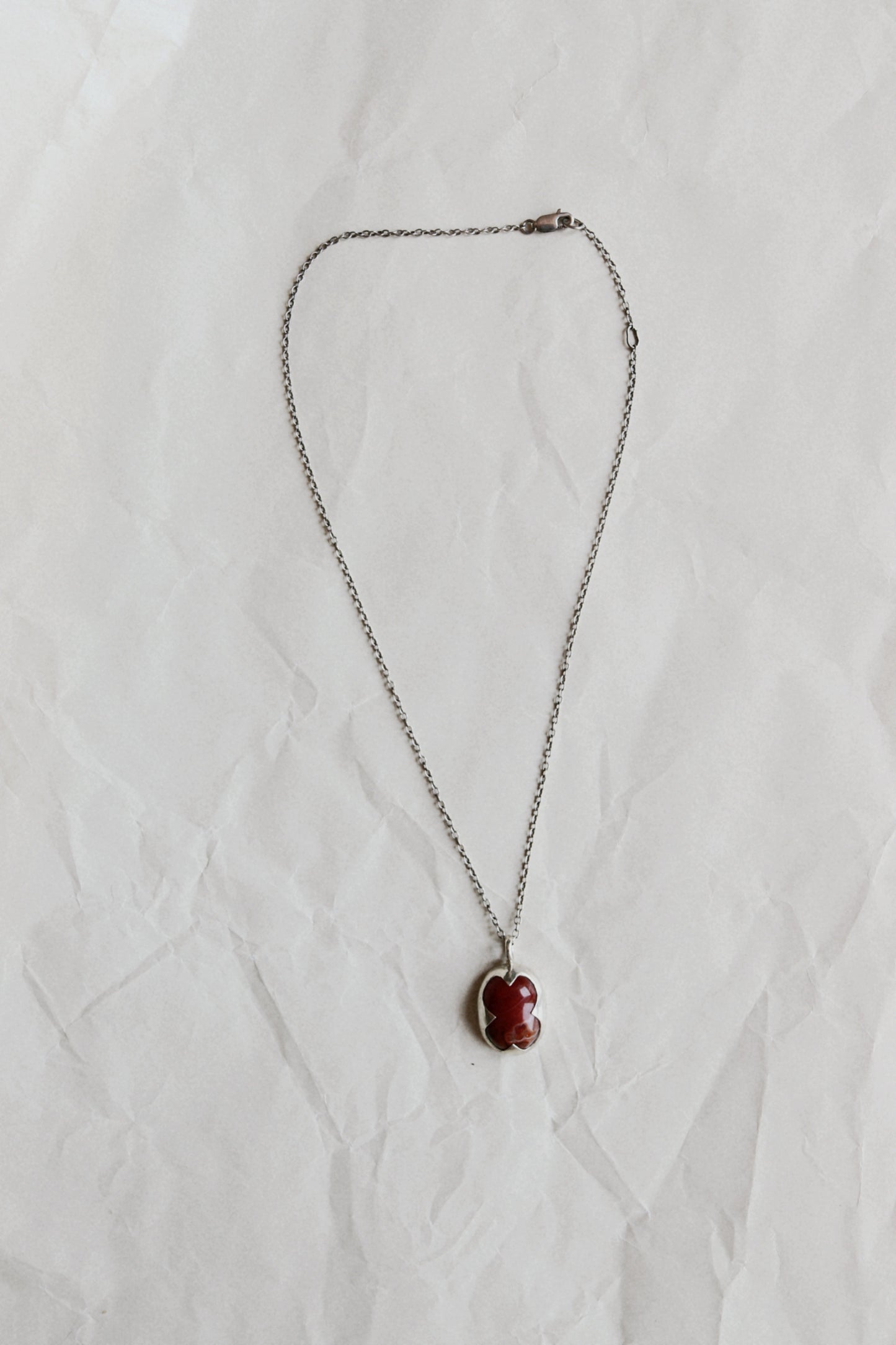 A silver chain necklace with an oval pendant featuring a carved red stone, laid flat on a wrinkled white paper background.