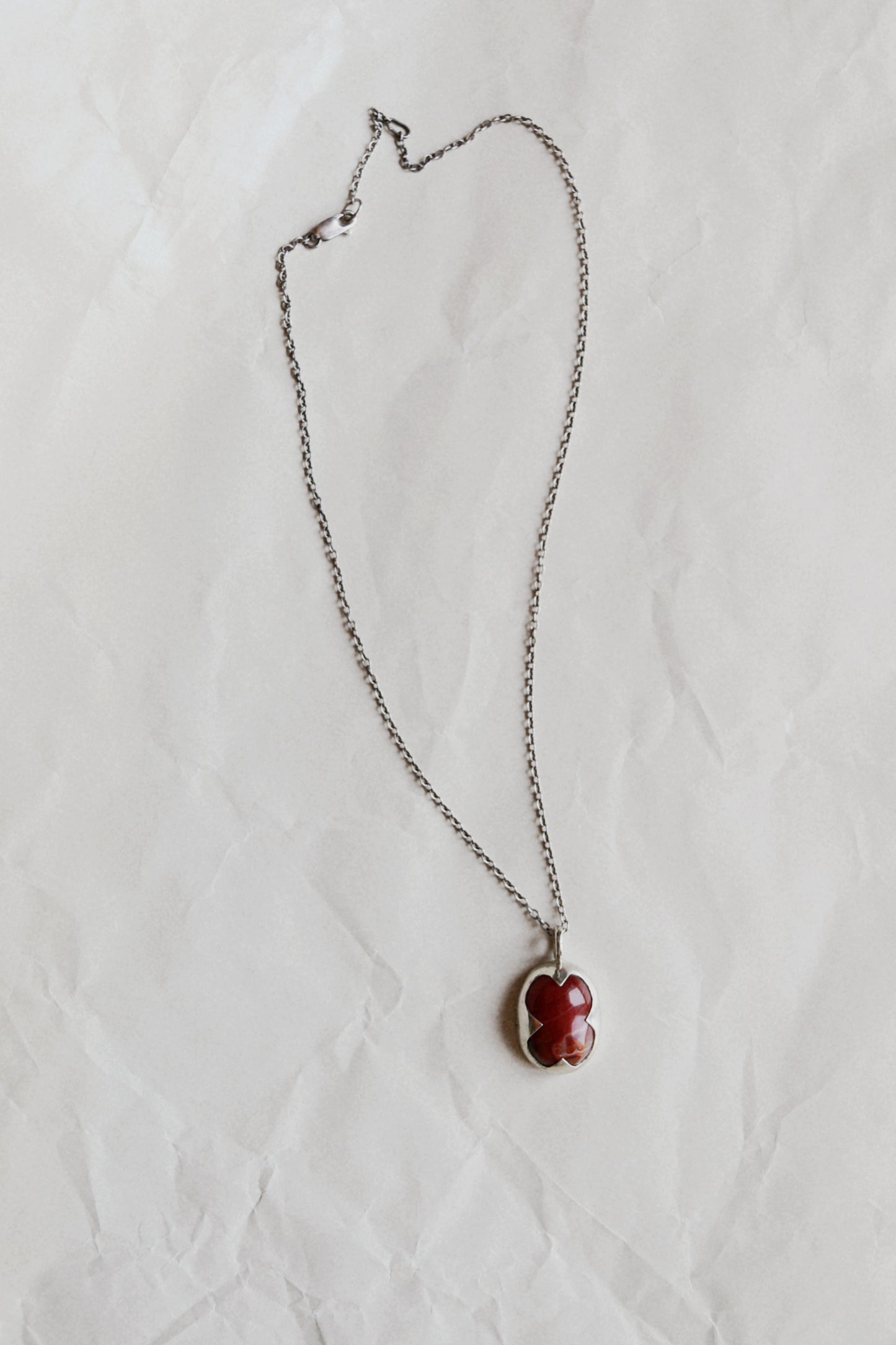 A silver chain necklace with a small, irregularly shaped red stone pendant, laid flat on a textured white paper background.