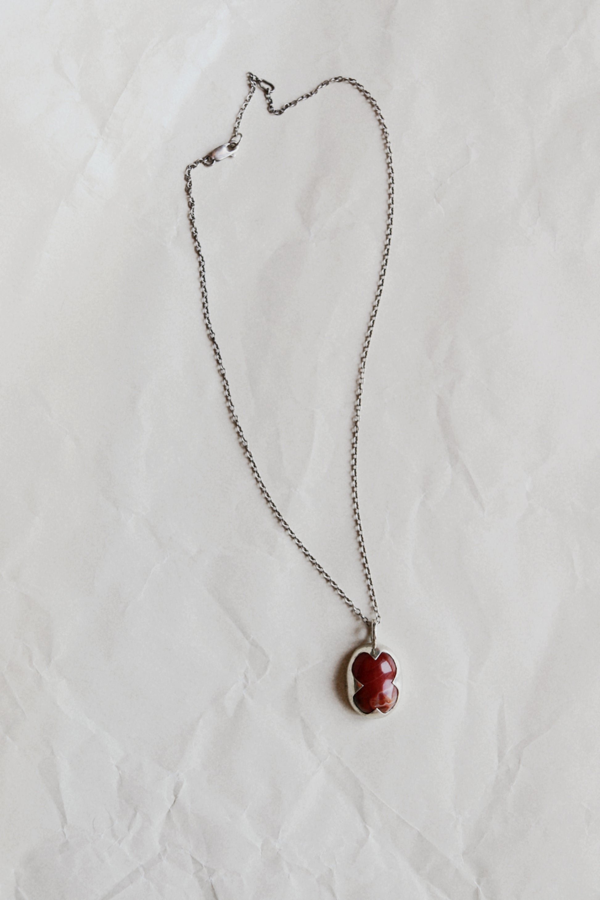 A silver chain necklace with a small, irregularly shaped red stone pendant, laid flat on a textured white paper background.