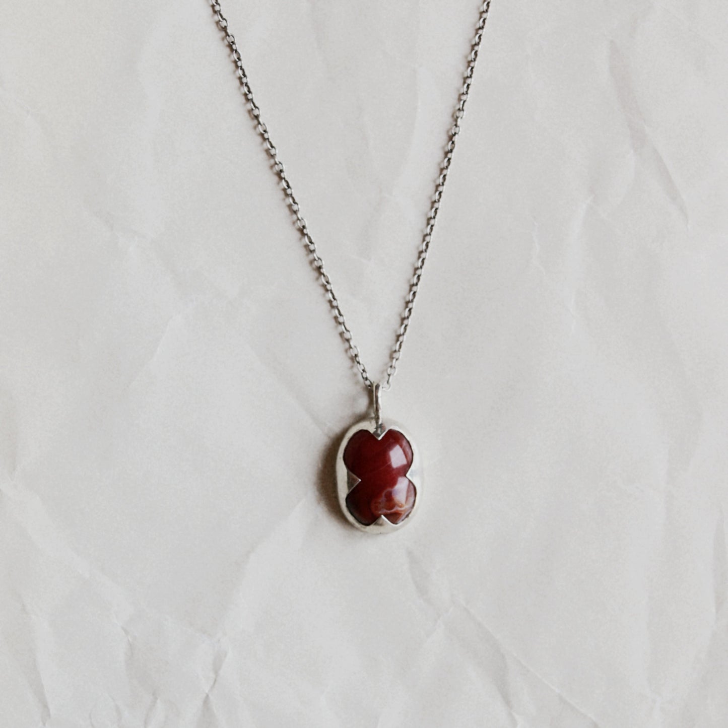 A silver chain necklace with a small clover-shaped pendant featuring a deep red center, displayed on a textured white background.