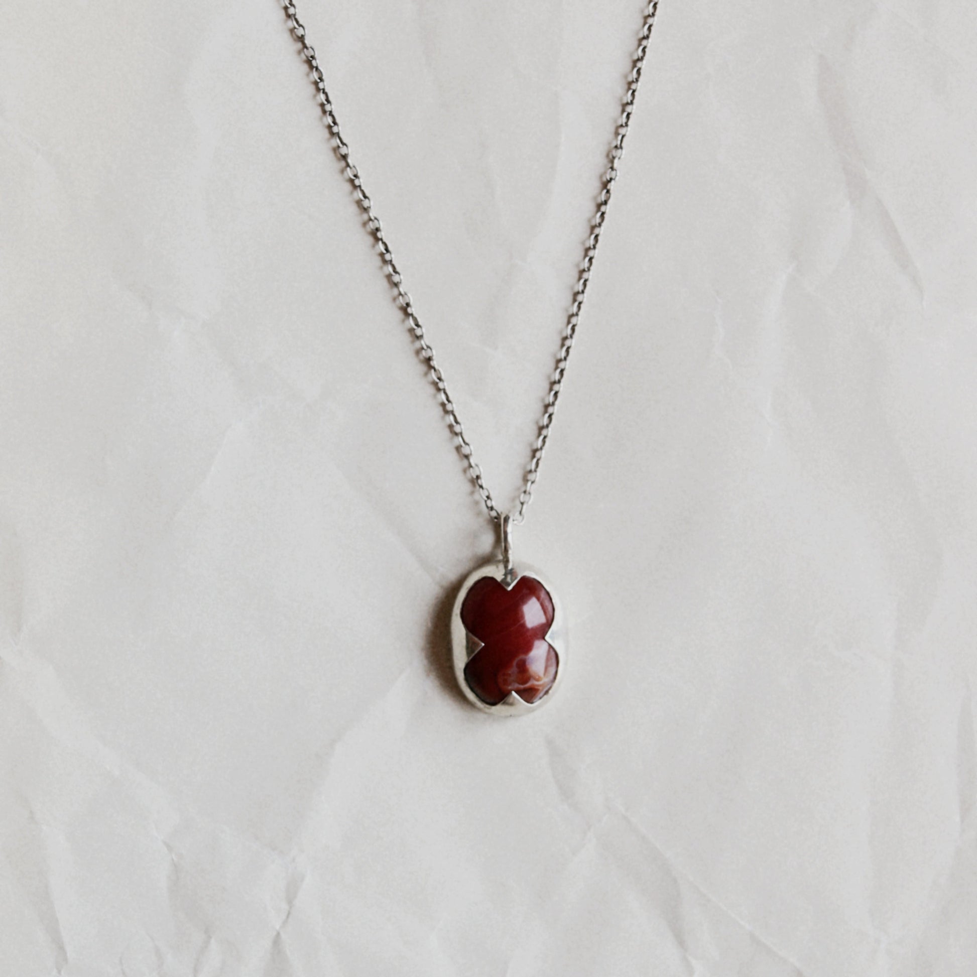 A silver chain necklace with a small clover-shaped pendant featuring a deep red center, displayed on a textured white background.
