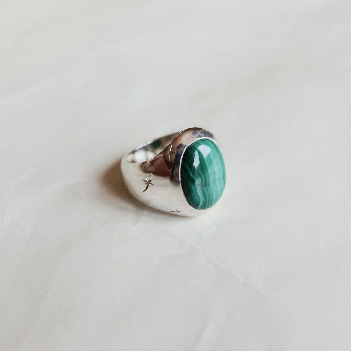 A silver ring featuring an oval green malachite stone, resting on a light beige textured surface.