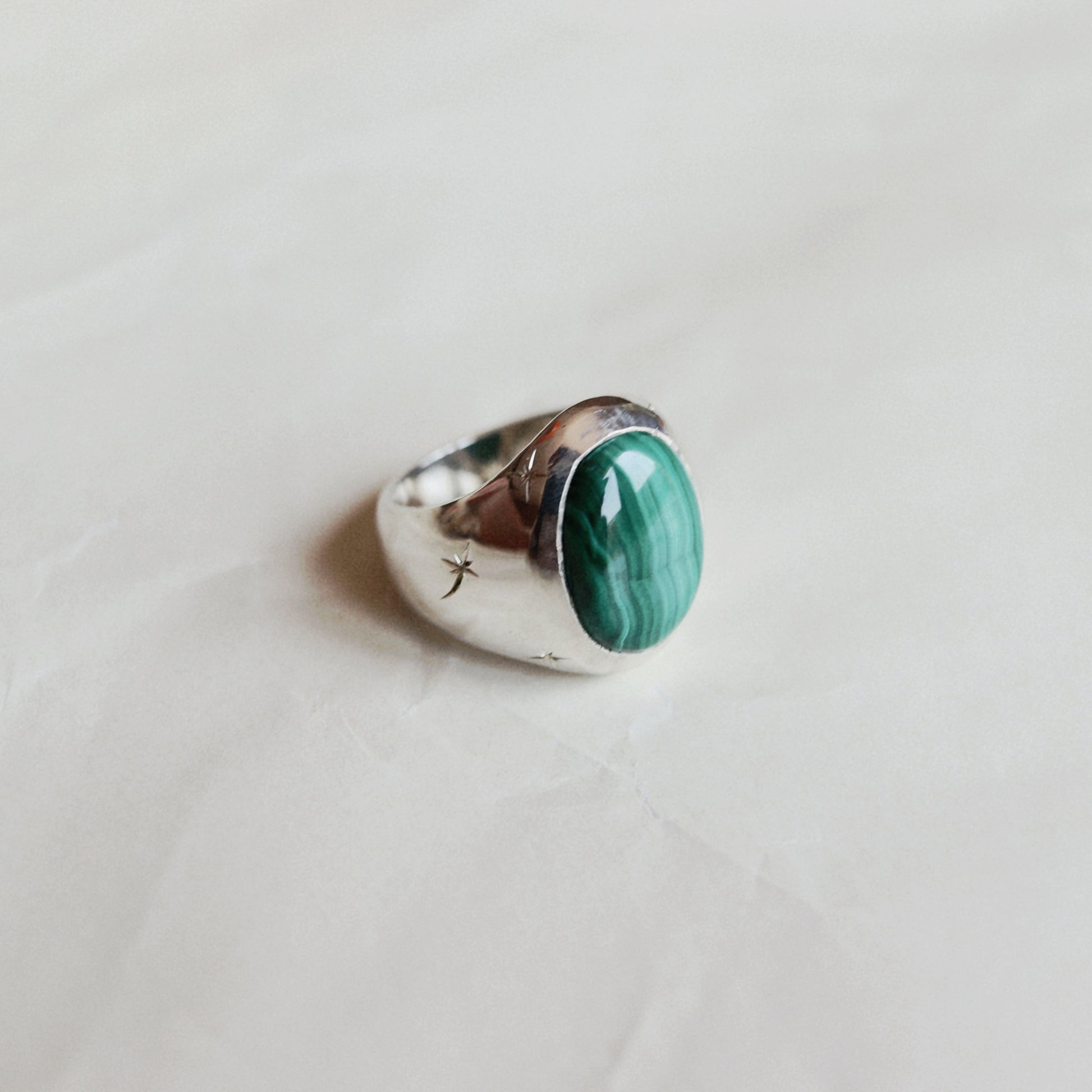 A silver ring featuring an oval green malachite stone, resting on a light beige textured surface.