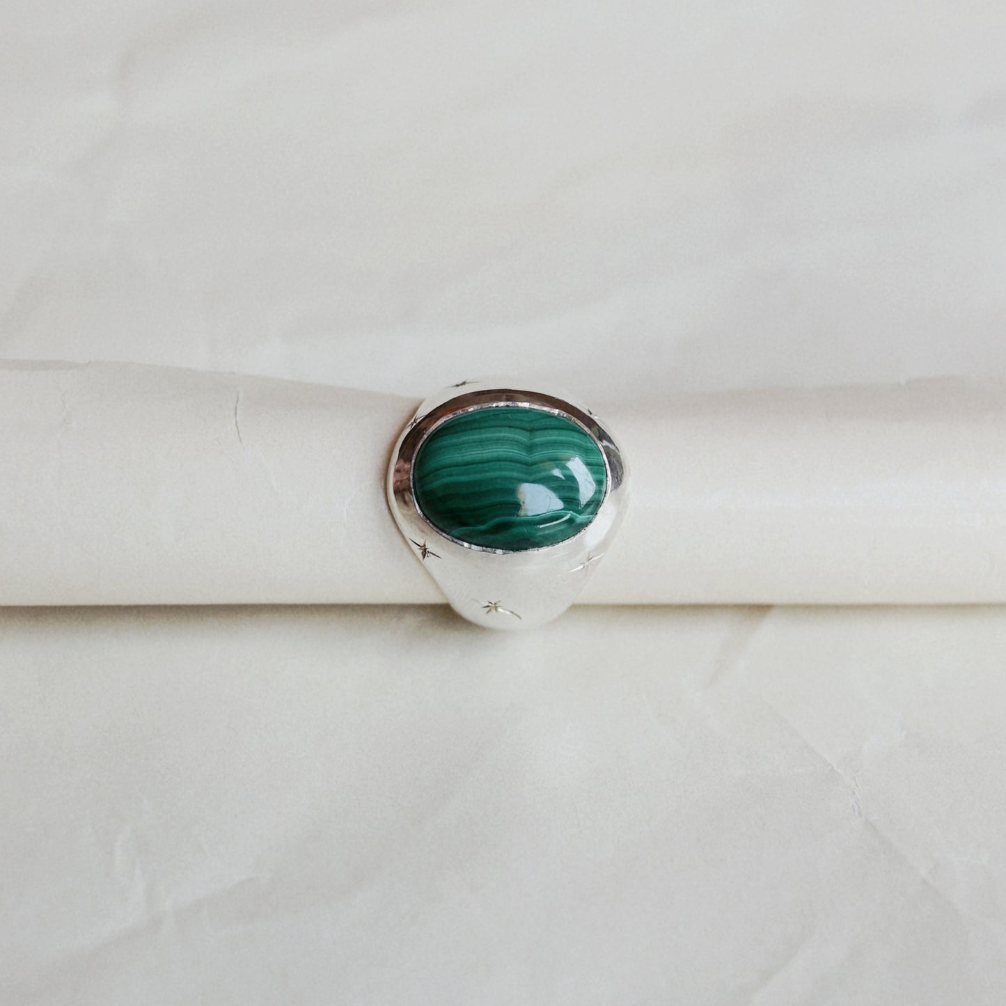 A silver ring featuring a round green malachite stone, displayed on a white paper ring holder against a textured white background.