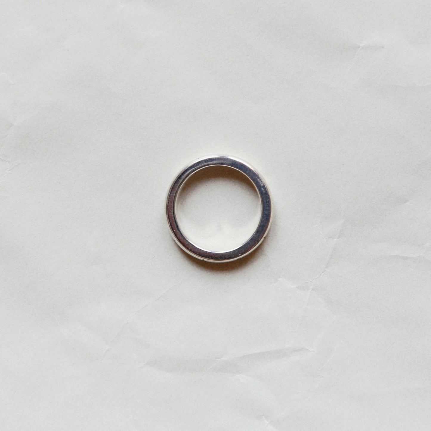 A simple silver-toned metal ring band photographed from above on a wrinkled off-white paper background.