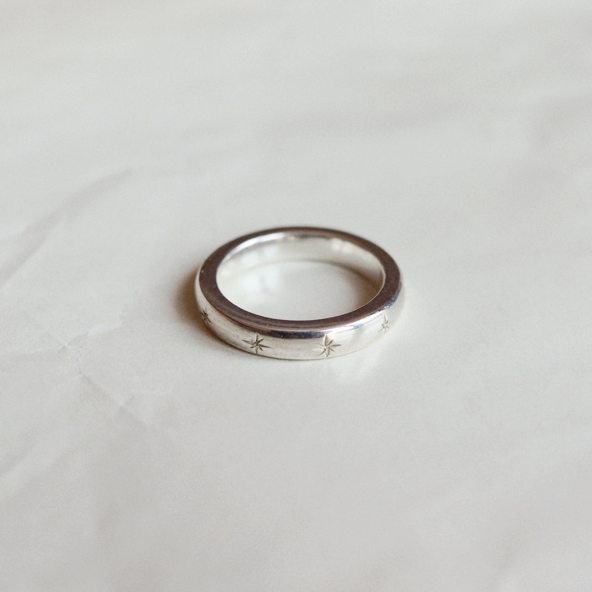 A thin silver band ring with small engraved star motifs on the front, resting on a light, slightly textured surface.