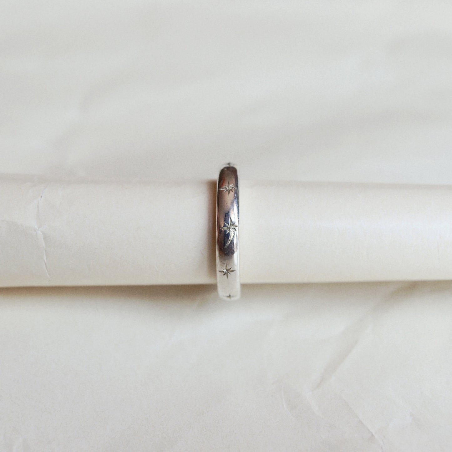 A slim silver band ring with engraved floral details, displayed on a white cylindrical ring stand against a soft, textured off-white background.