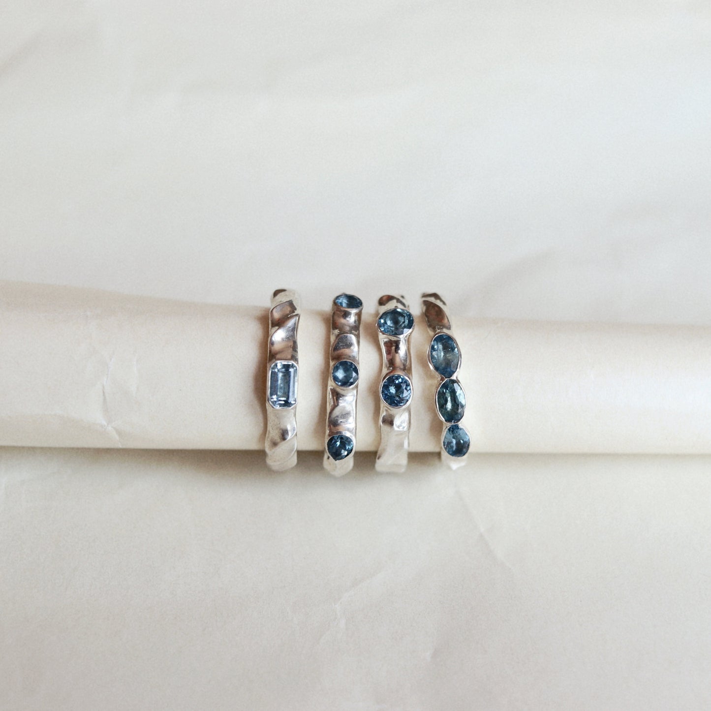 Four silver rings with blue gemstones displayed on a cream-colored ring stand against a light beige background.