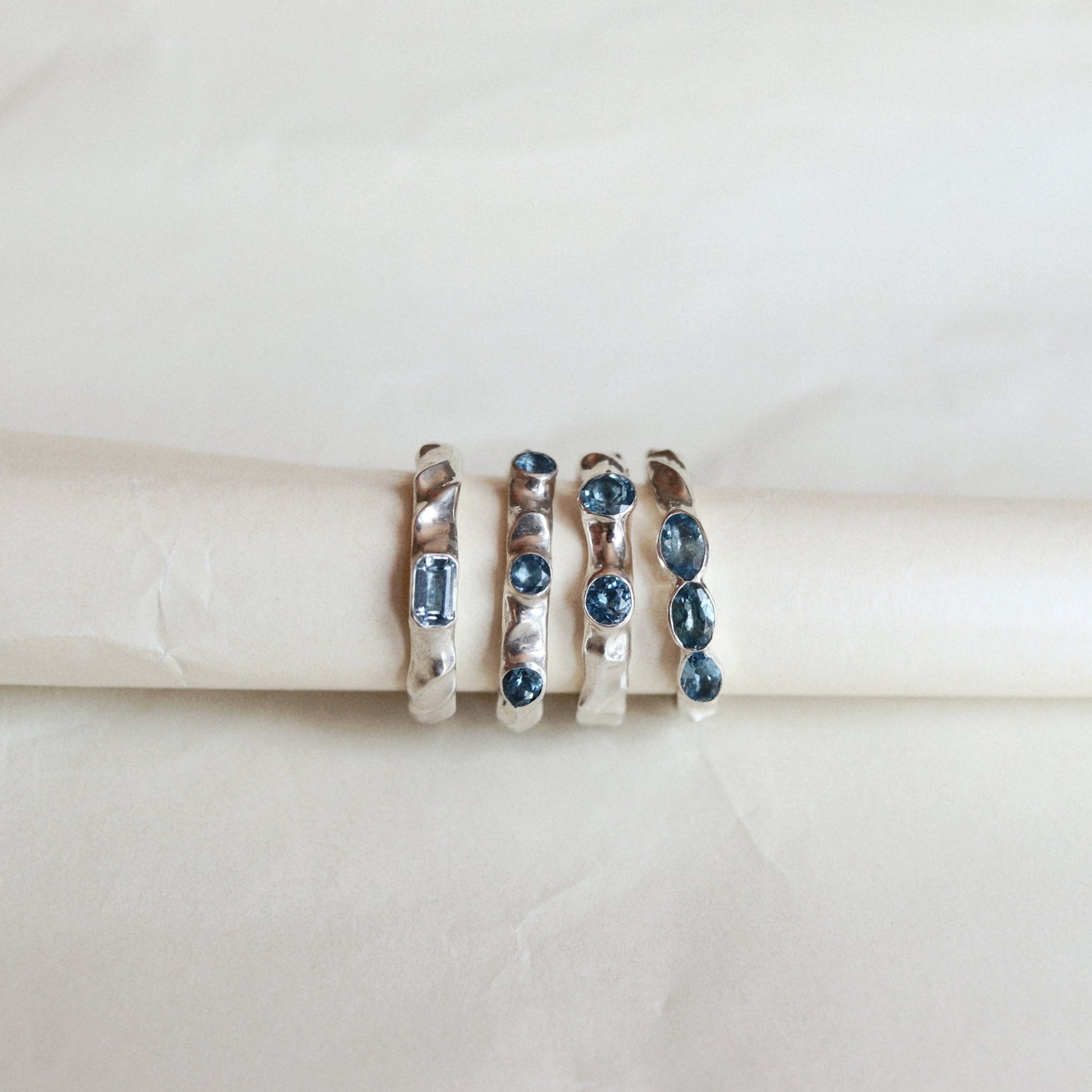 Four silver rings with blue gemstones displayed on a cream-colored ring stand against a light beige background.