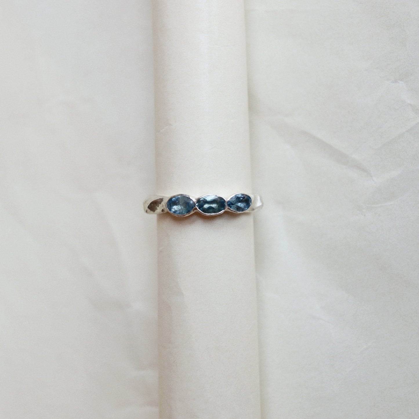 A silver ring featuring three small, oval-cut dark blue gemstones set vertically, displayed on a textured off-white surface.