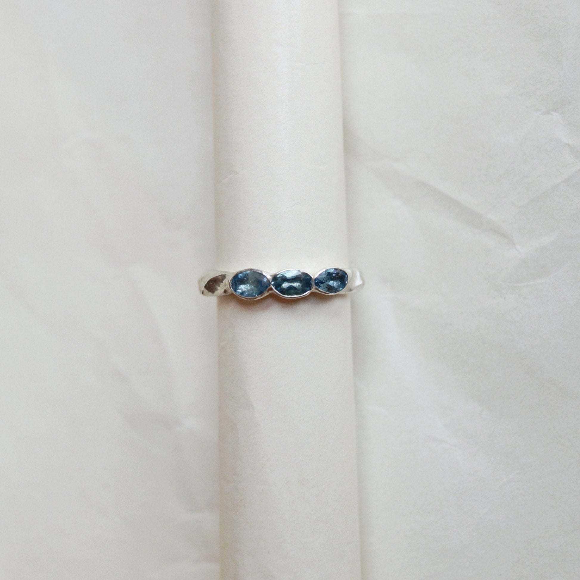 A silver ring featuring three small, oval-cut dark blue gemstones set vertically, displayed on a textured off-white surface.