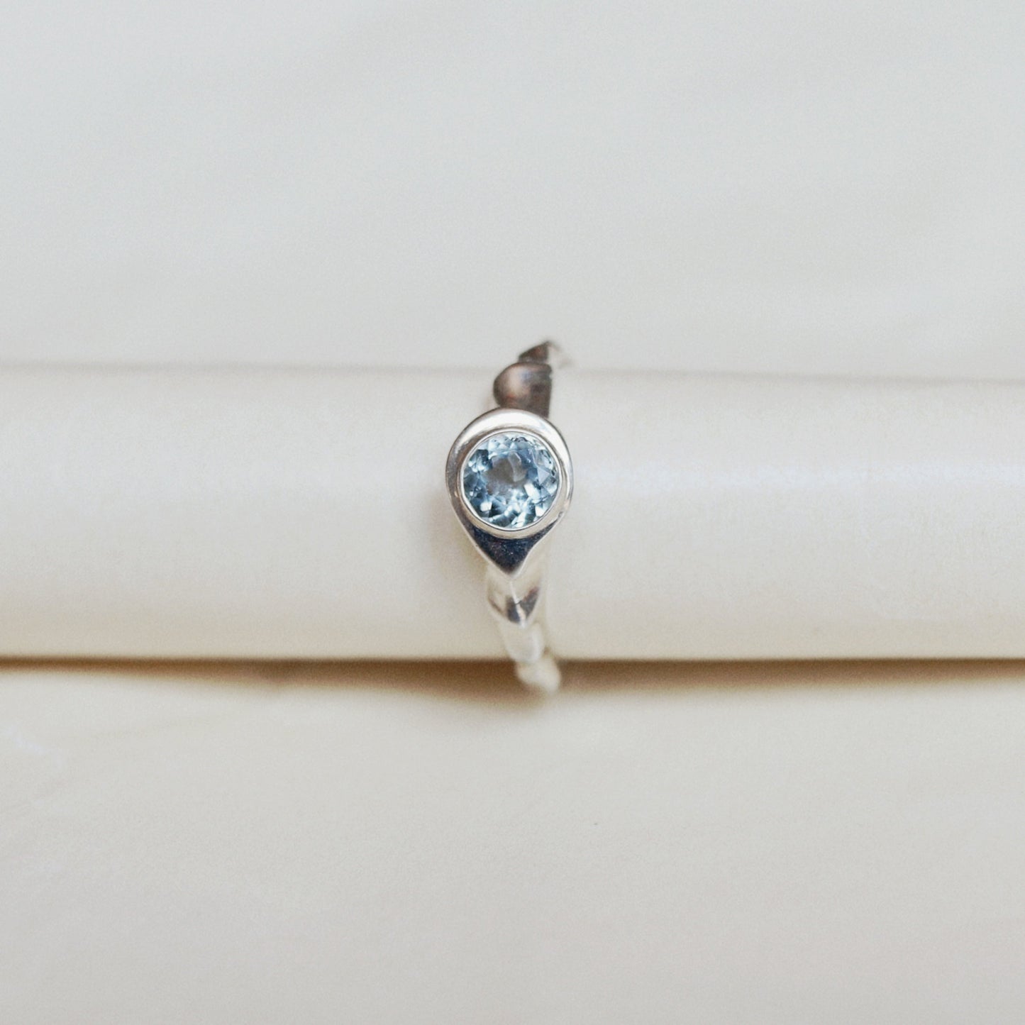 A silver ring featuring a round-cut light blue gemstone, displayed on a cream-colored ring stand.