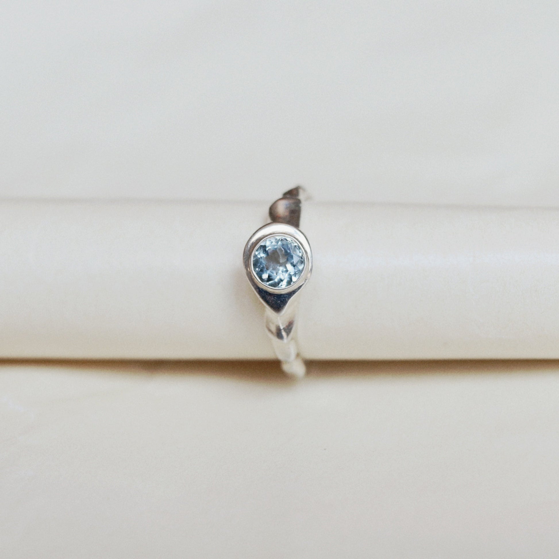 A silver ring featuring a round-cut light blue gemstone, displayed on a cream-colored ring stand.