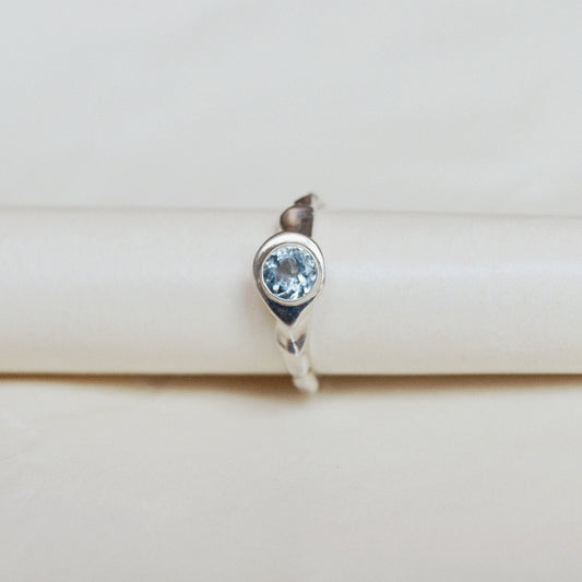 A silver ring featuring a round-cut light blue gemstone, displayed on a cream-colored ring stand.