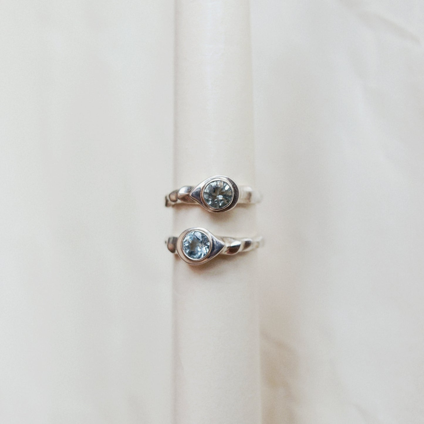Two silver-toned rings with oval-cut clear gemstones displayed on a cream-colored ring stand against a soft beige fabric background.