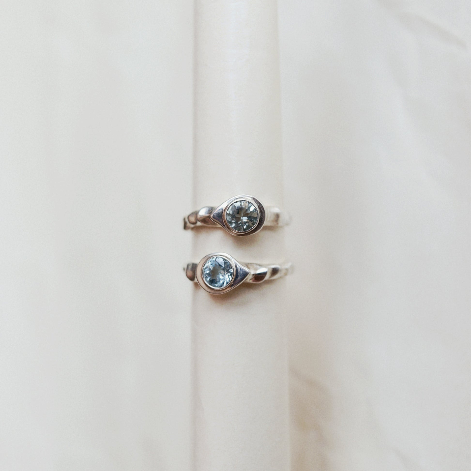 Two silver-toned rings with oval-cut clear gemstones displayed on a cream-colored ring stand against a soft beige fabric background.