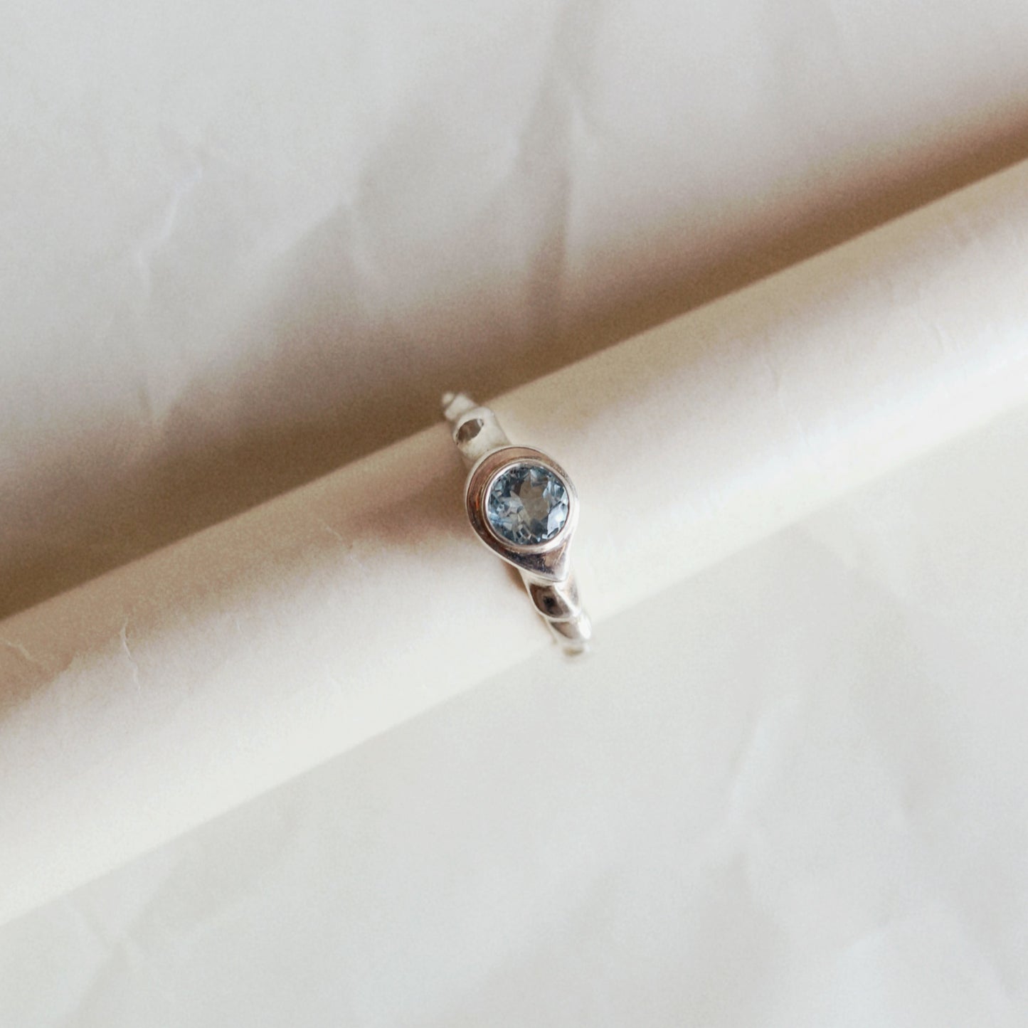 A silver ring with a round blue gemstone, displayed on a rolled cream-colored paper cylinder against a matching textured paper background.