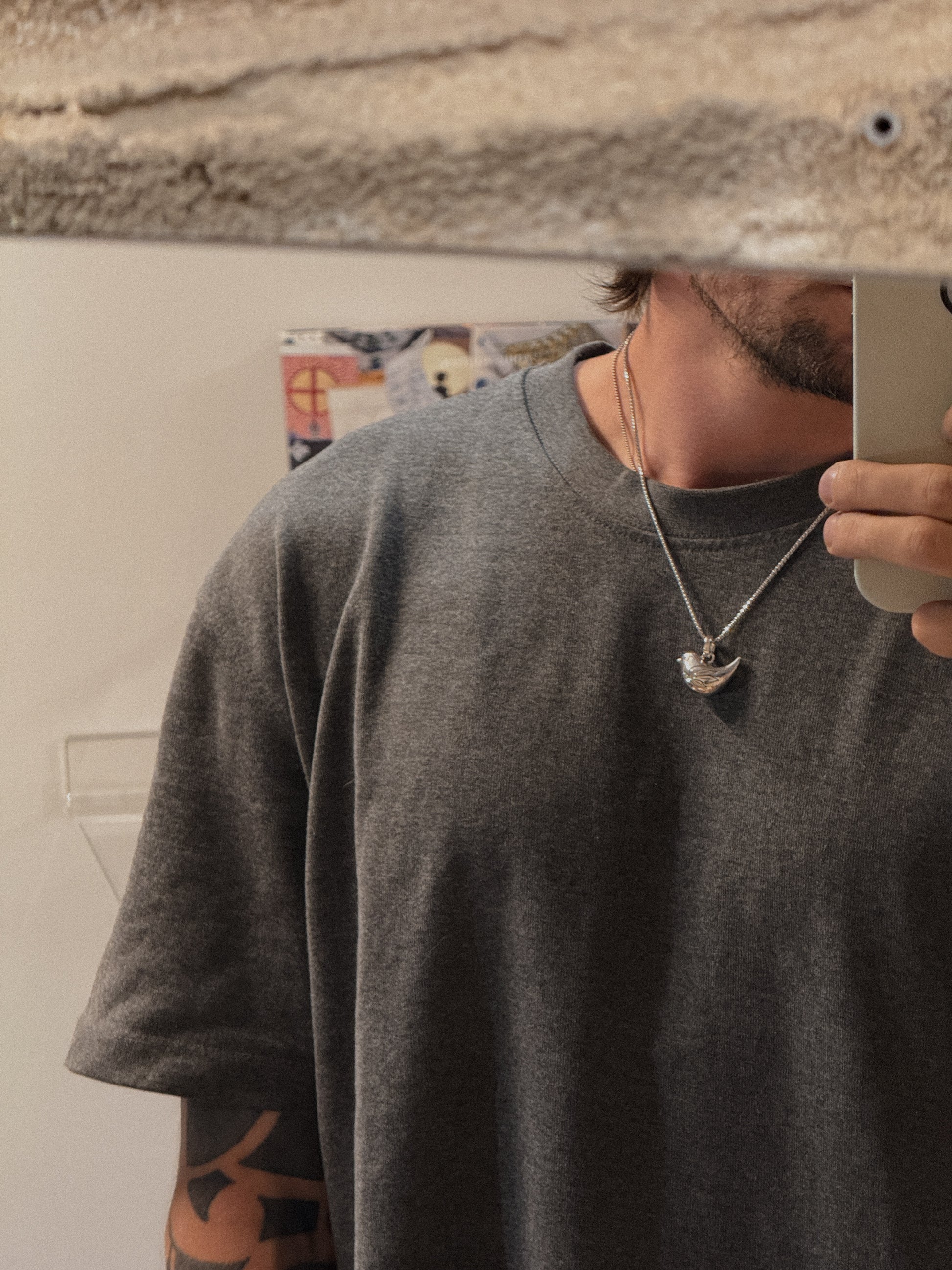 Person wearing a gray t-shirt and a necklace with a heart pendant, taking a mirror selfie.