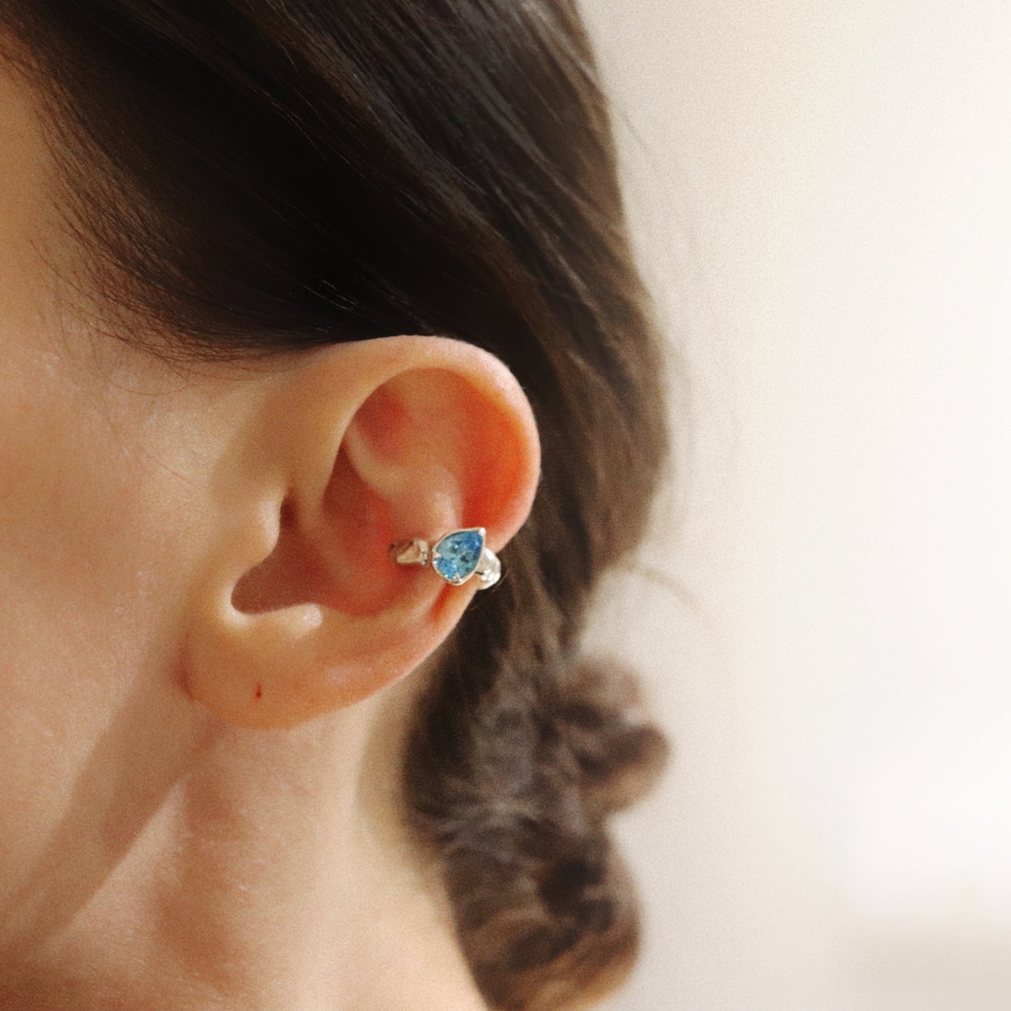 A close-up side profile of a person's ear wearing a small gold ear cuff with a blue heart-shaped gemstone.