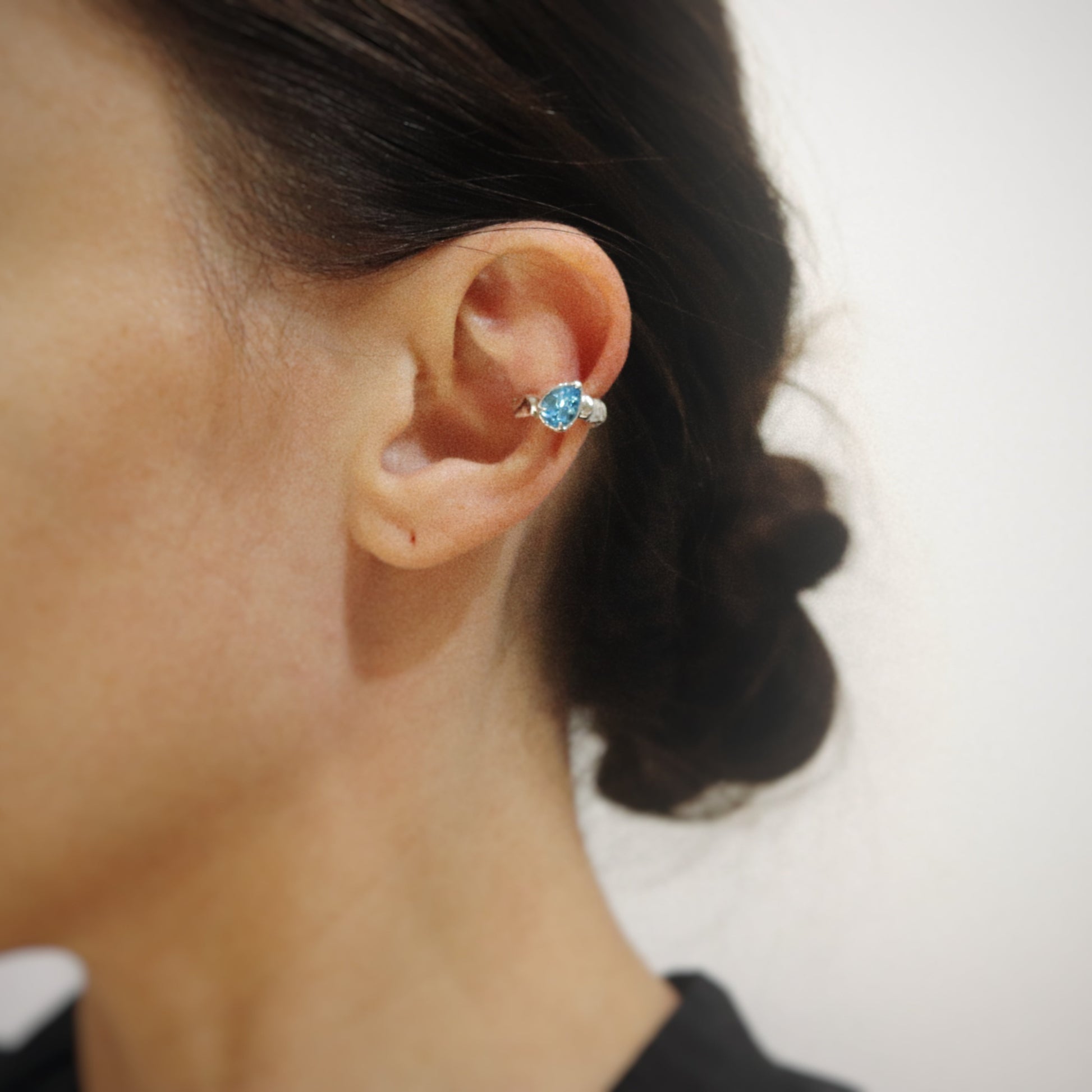 A close-up of a person's ear wearing a silver-toned ear cuff with a prominent oval light blue gemstone.