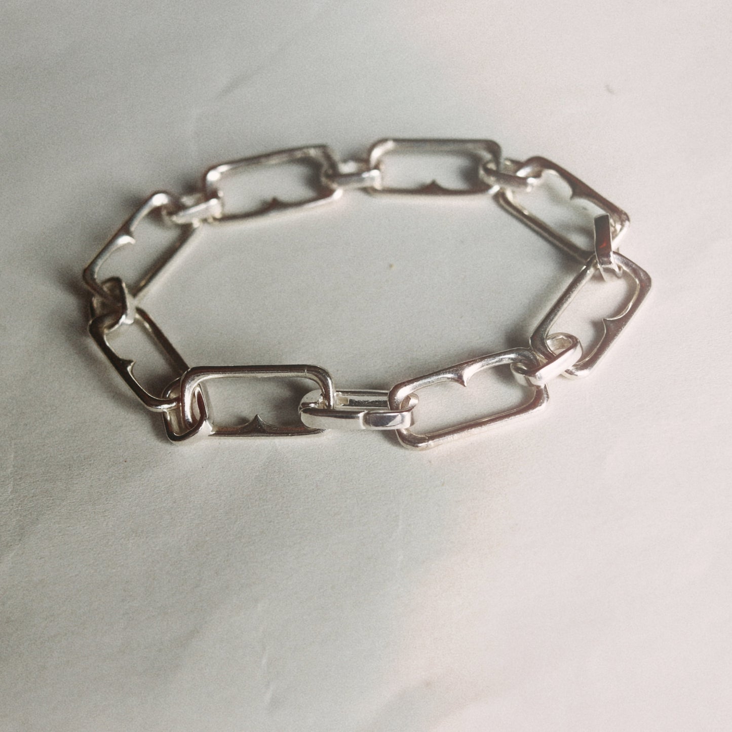 A silver-toned metal link bracelet with elongated rectangular links, arranged in an oval shape on a light off-white background.