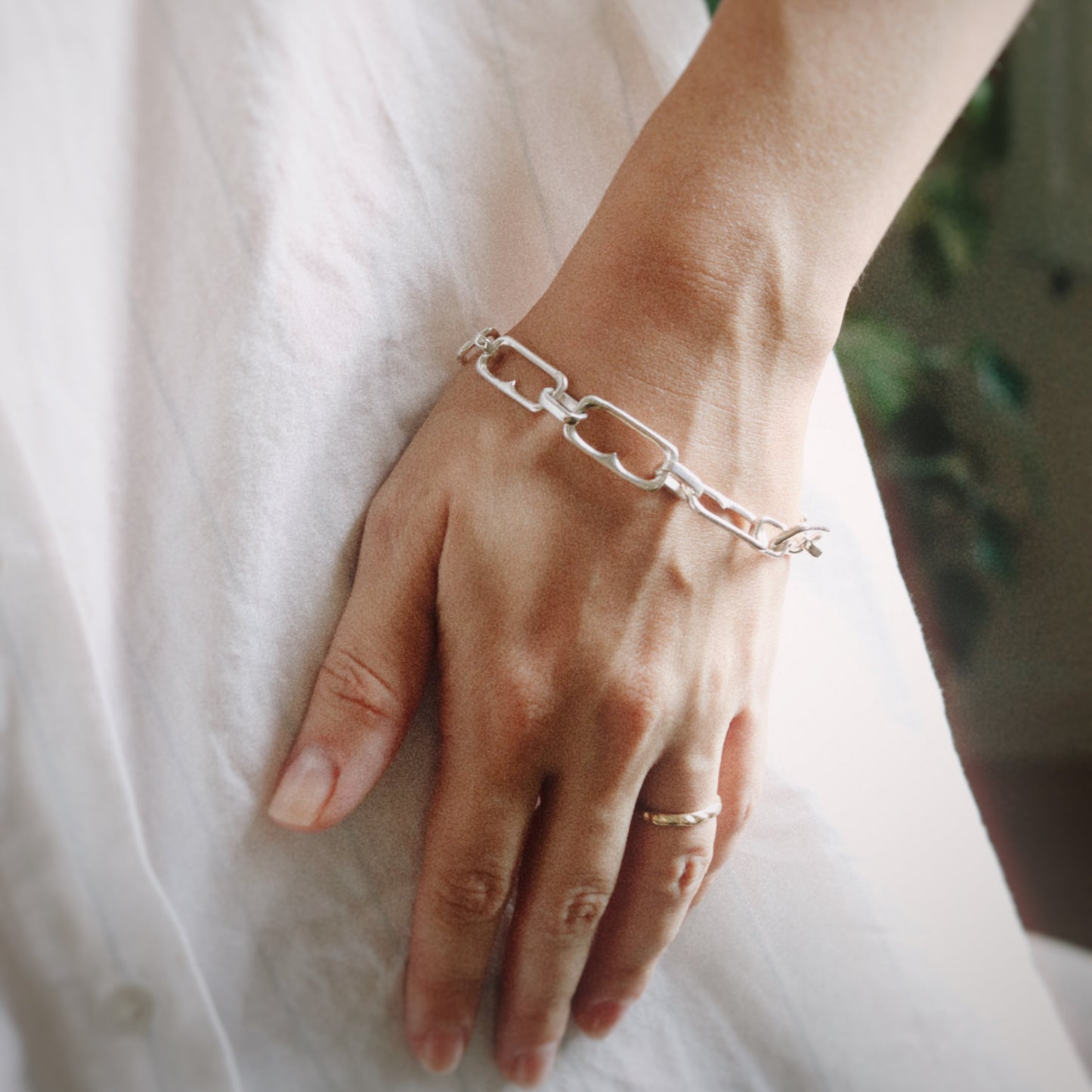 A close-up of a person's hand resting on a white textured fabric, wearing a delicate silver chain link bracelet and a thin silver ring.