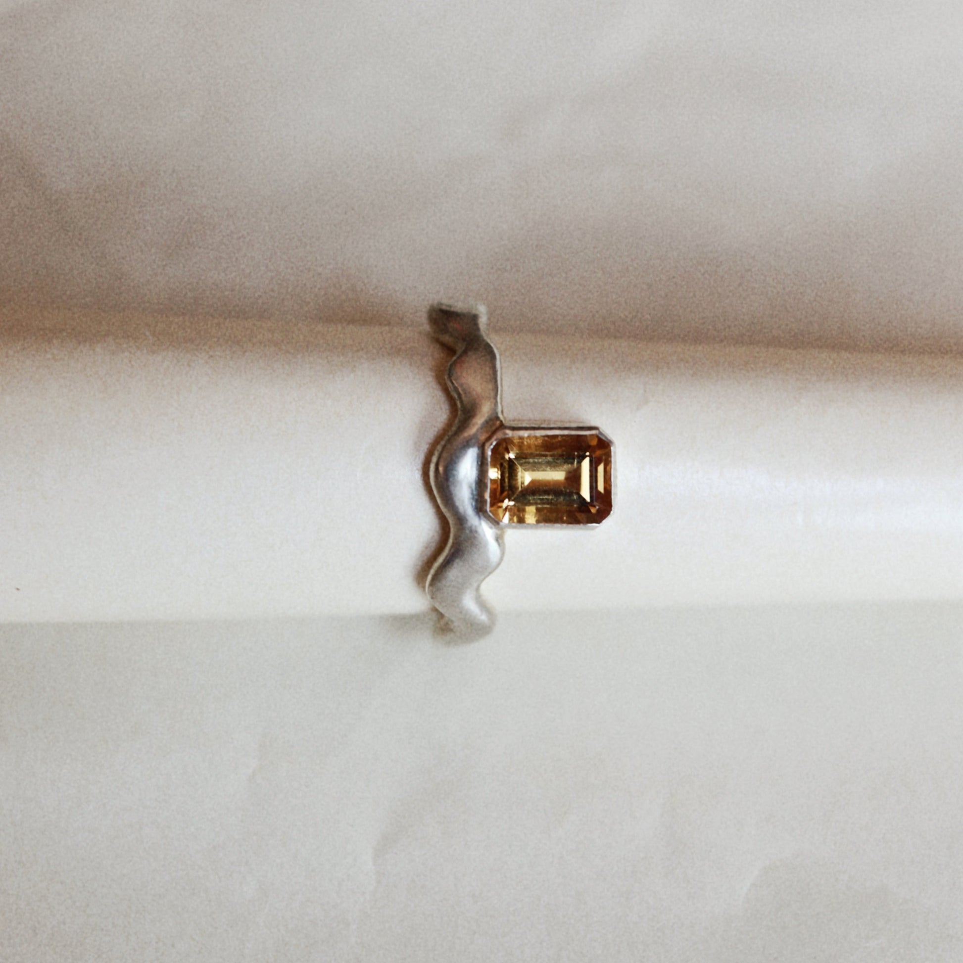 A silver ring with a wavy band and a rectangular yellow gemstone, shown resting on a white cylindrical surface.