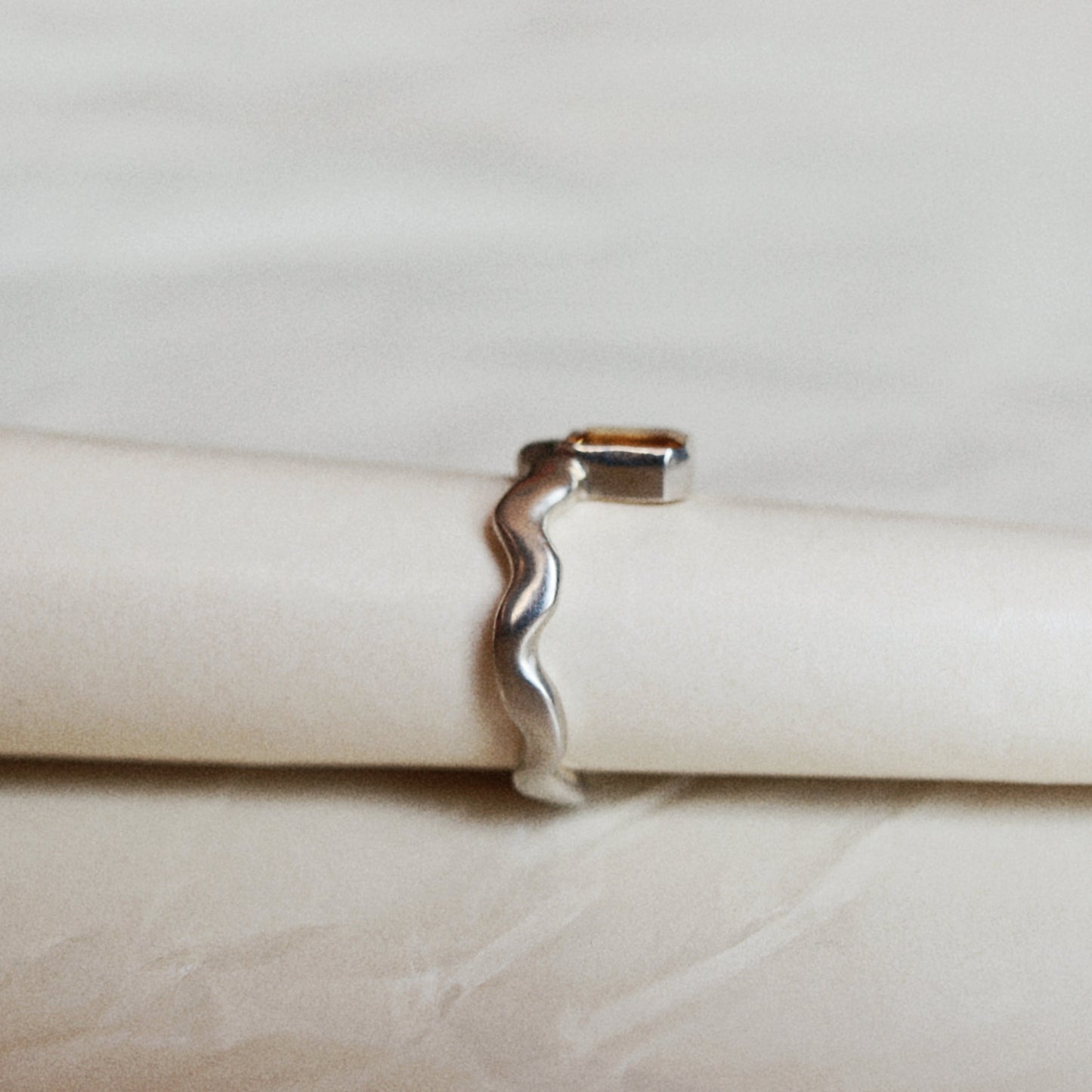 A silver wavy open band ring displayed on a cream-colored ring holder against a light beige background.