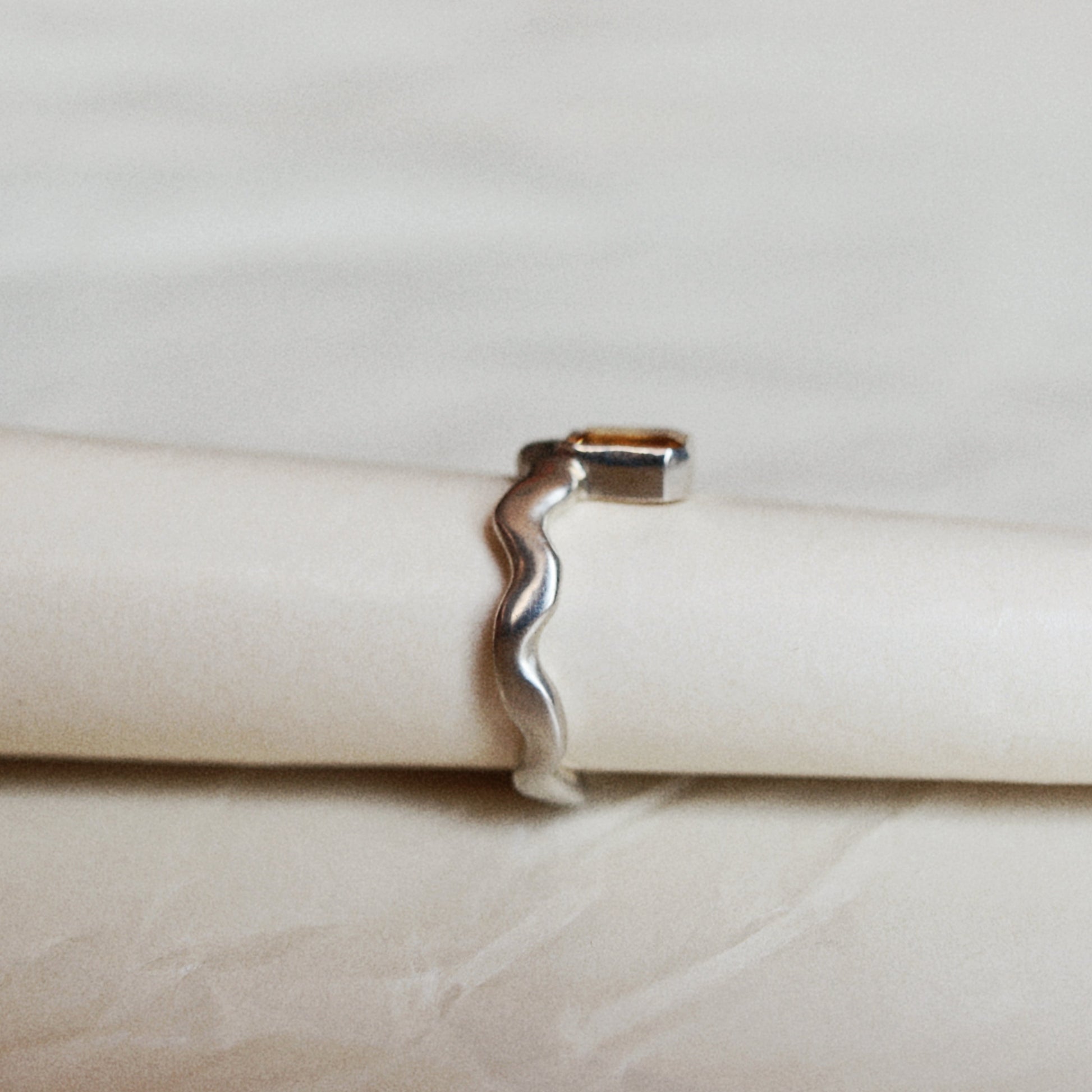 A silver wavy open band ring displayed on a cream-colored ring holder against a light beige background.