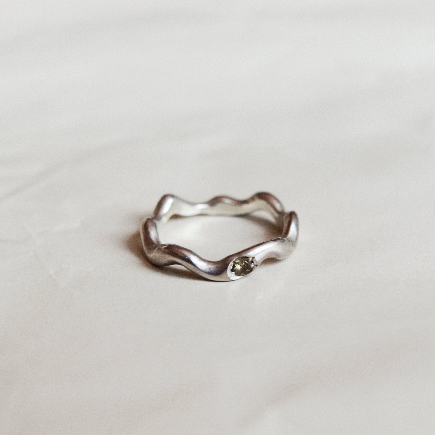A silver wavy band ring with a small round clear gemstone, resting on a light off-white textured surface.