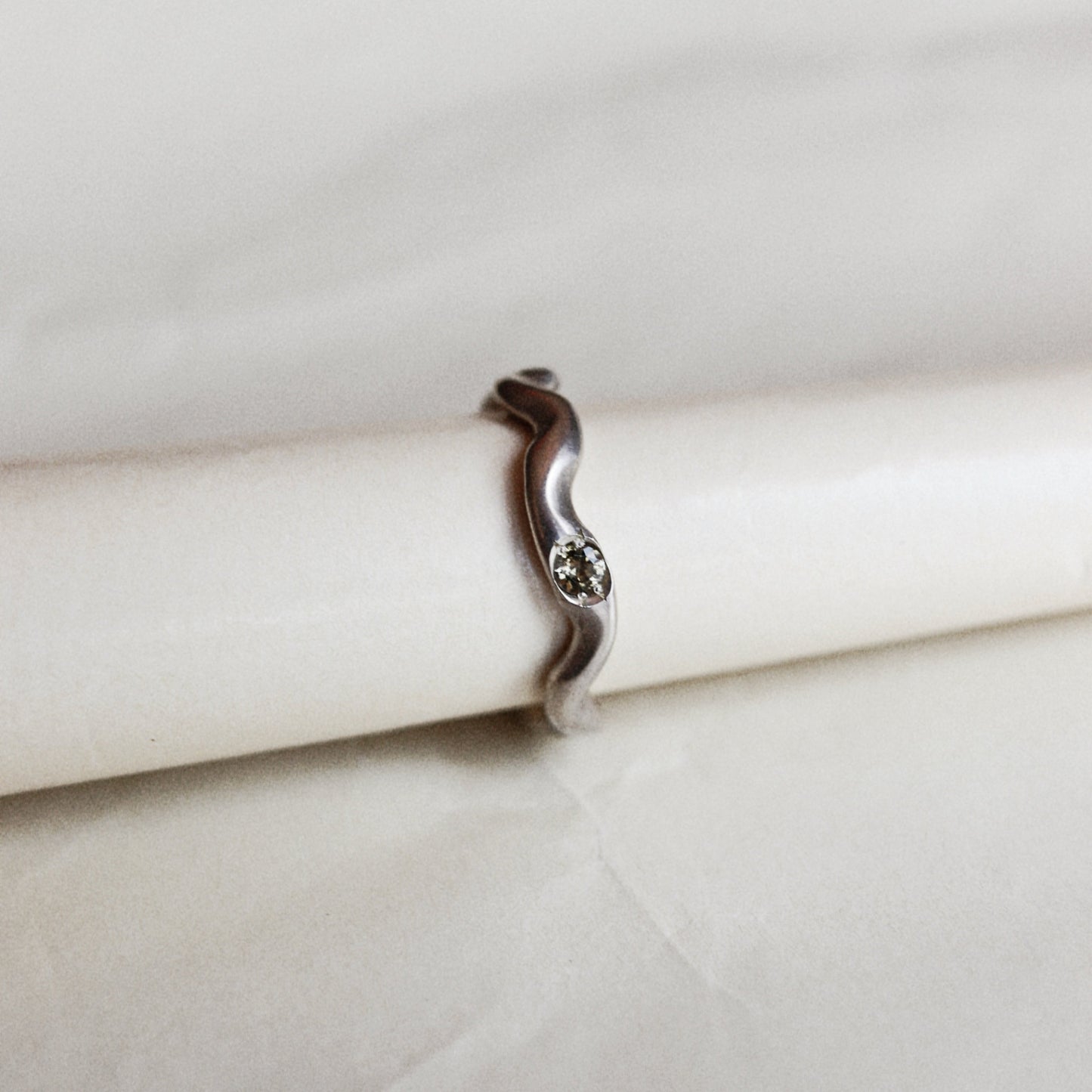 A silver wavy band ring with a small central clear gemstone, displayed on a white cylindrical stand against a light beige fabric background.