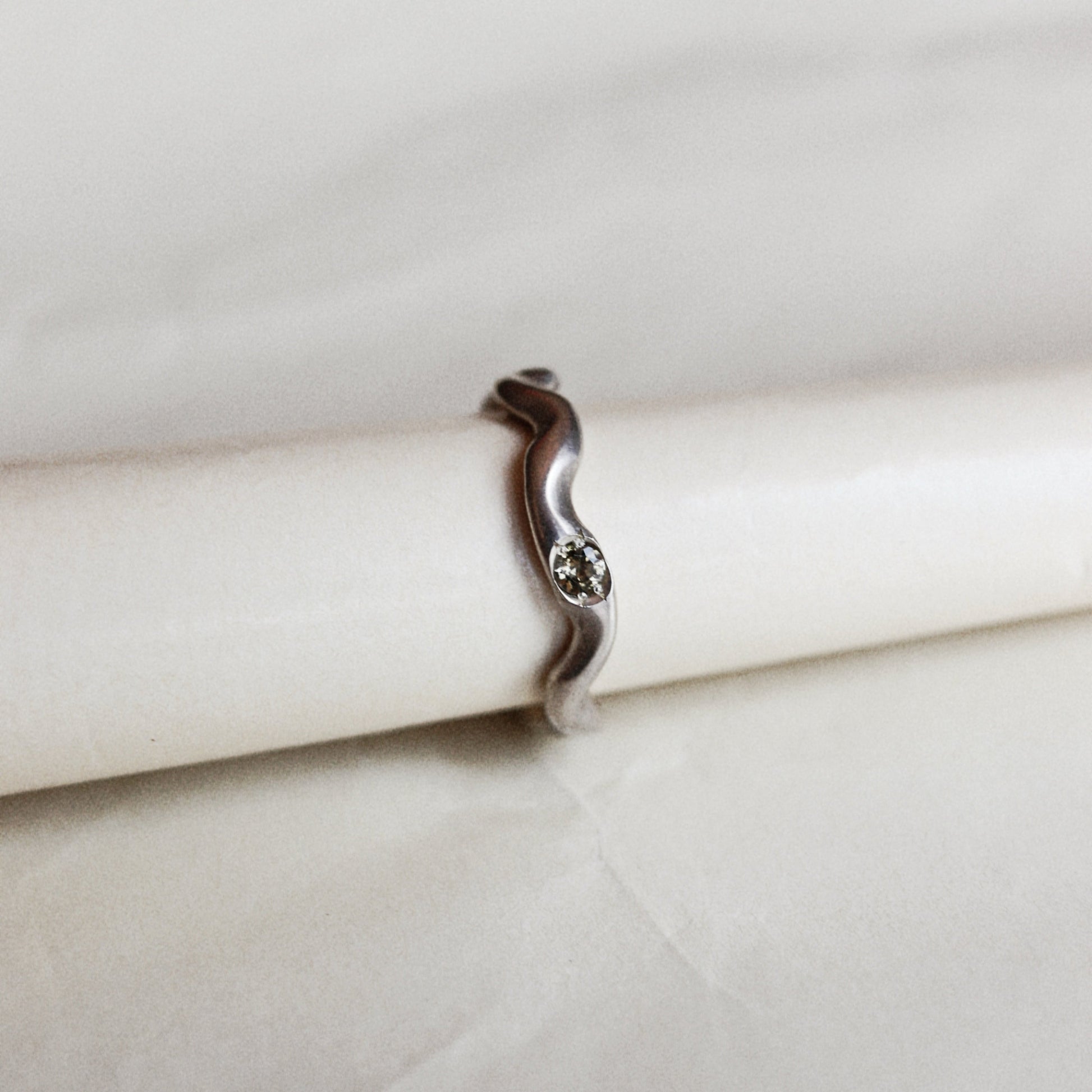 A silver wavy band ring with a small central clear gemstone, displayed on a white cylindrical stand against a light beige fabric background.