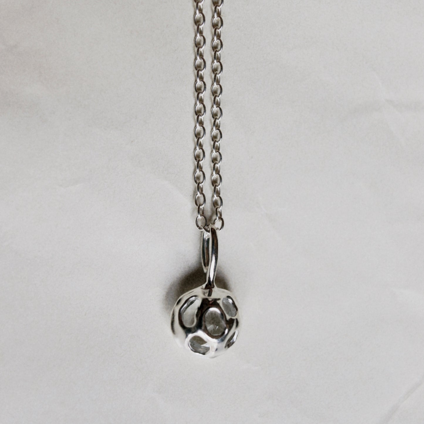A silver ball pendant necklace with a textured, faceted design hanging from a delicate silver chain against a wrinkled white paper background.