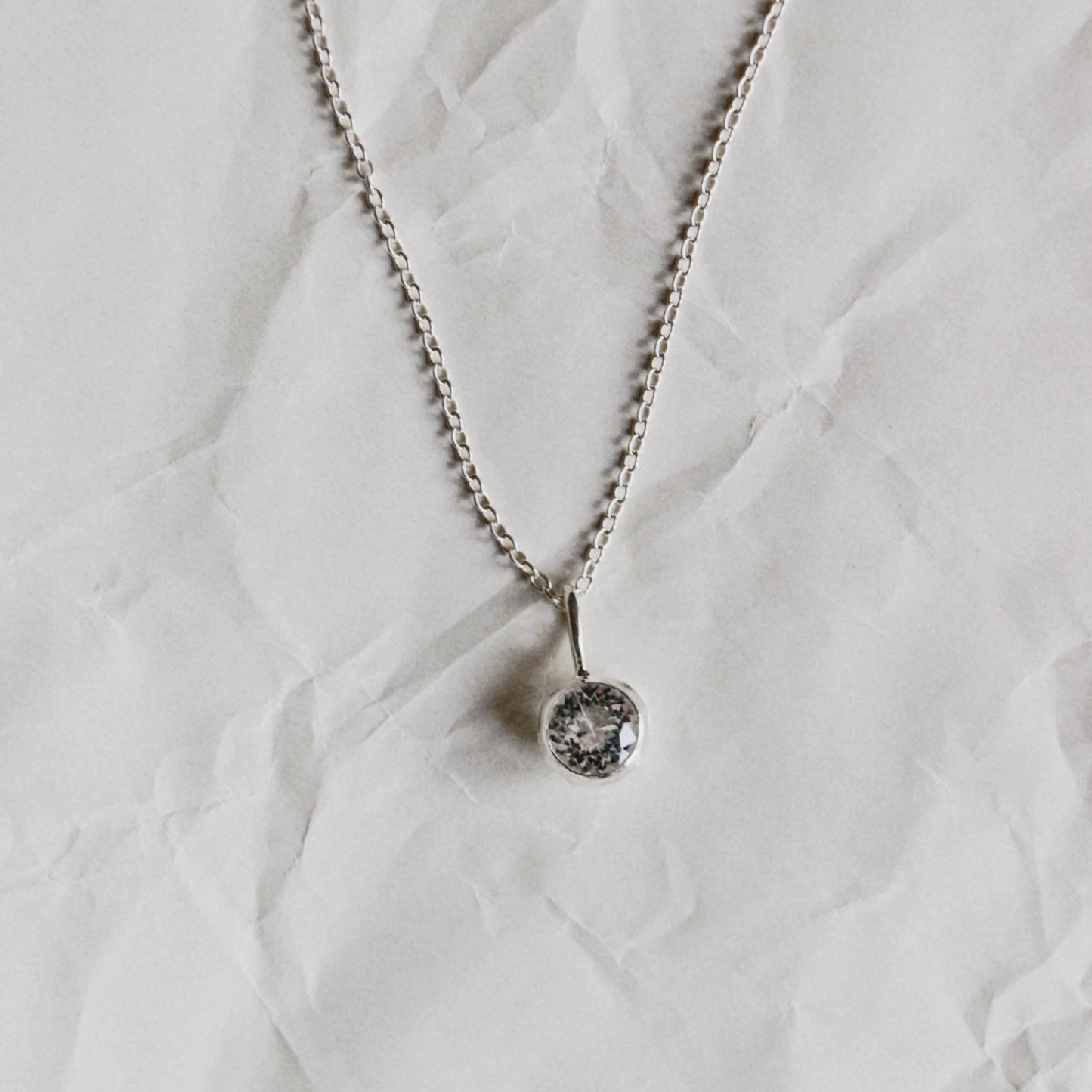 A silver chain necklace with a small, round bezel-set clear gemstone pendant, laid flat on a textured, crumpled white paper background.