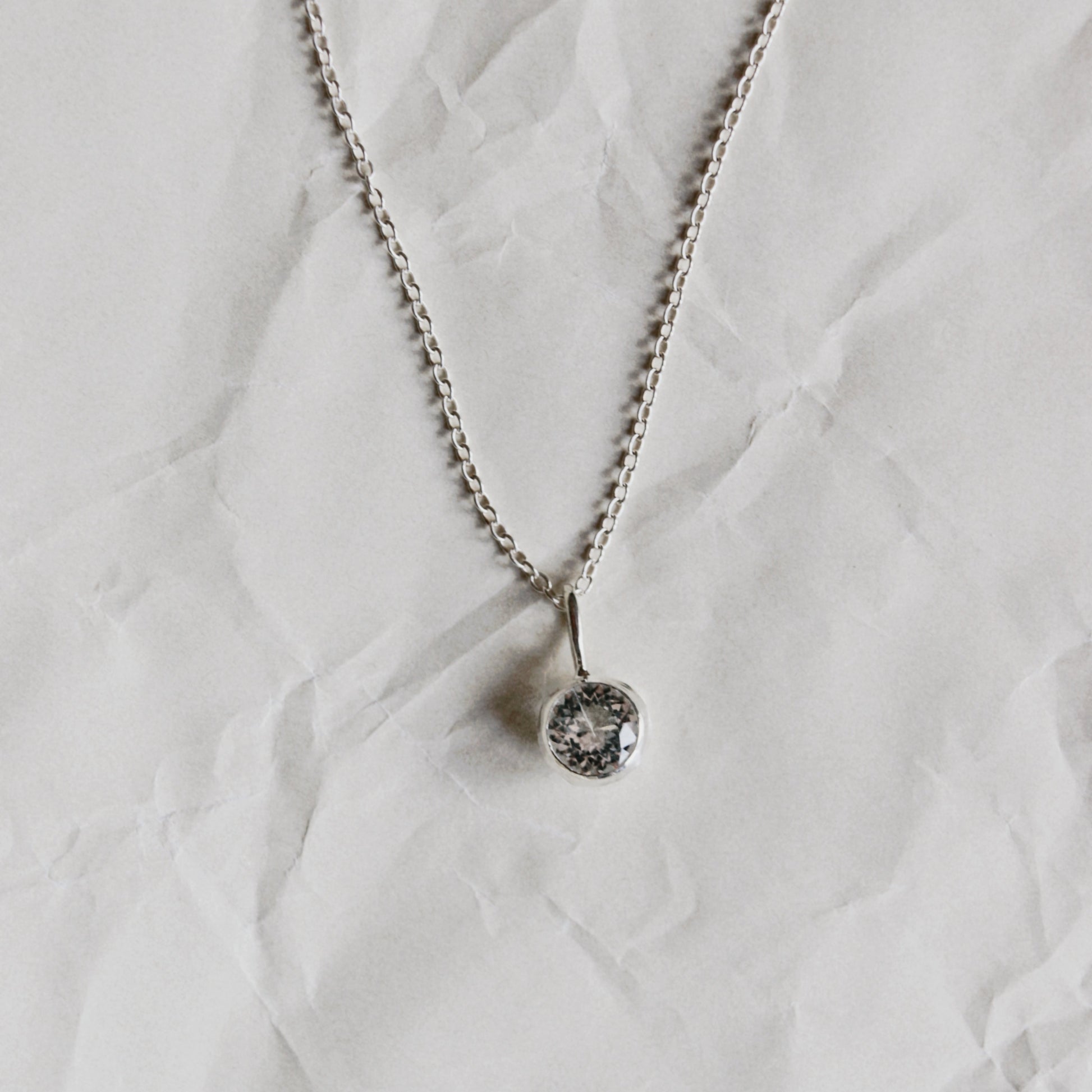 A silver chain necklace with a small, round bezel-set clear gemstone pendant, laid flat on a textured, crumpled white paper background.