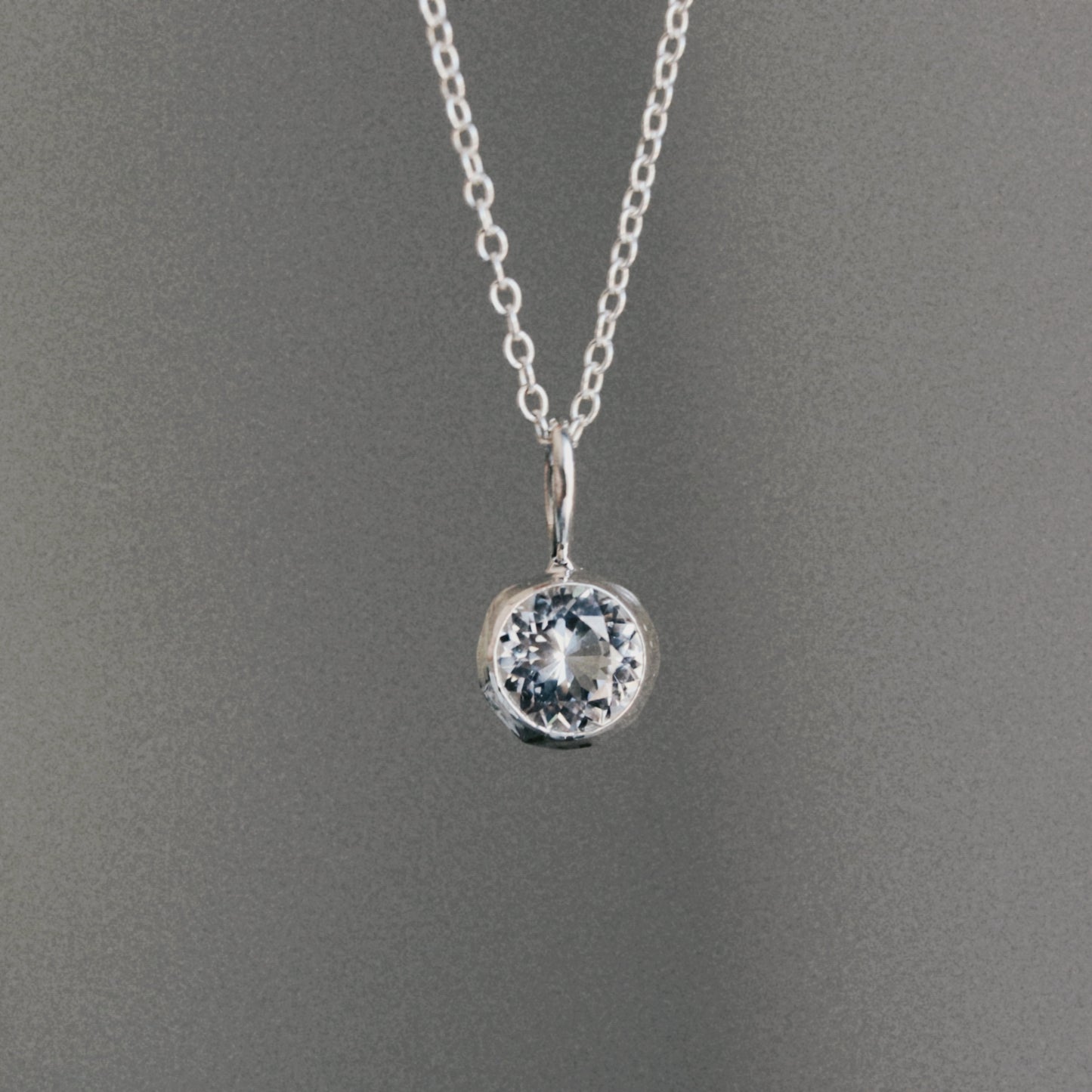 A silver chain necklace with a round bezel-set clear gemstone pendant, displayed against a neutral gray background.